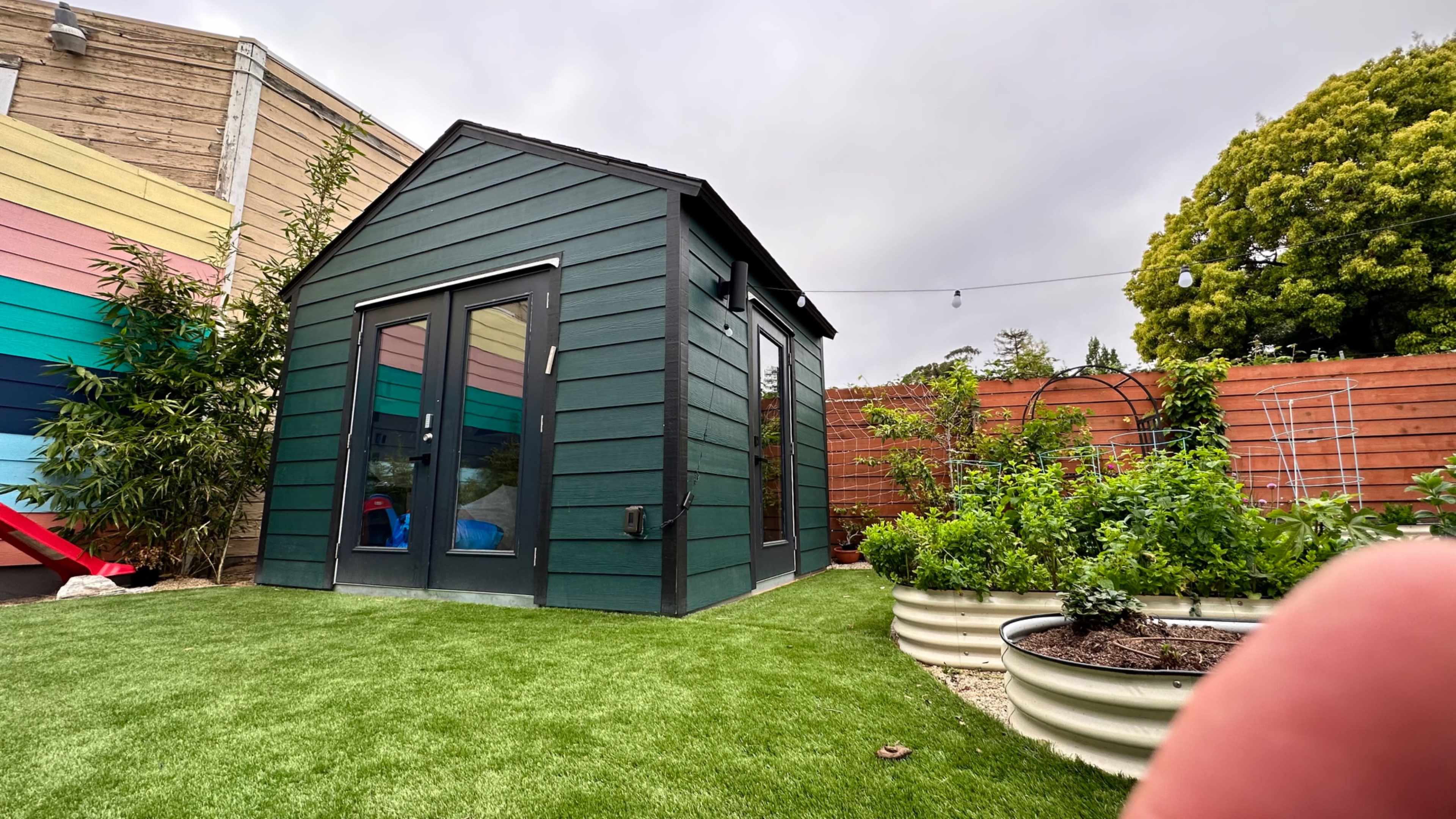 A small green shed with large windows is situated on a grassy area surrounded by garden beds and colorful fencing.