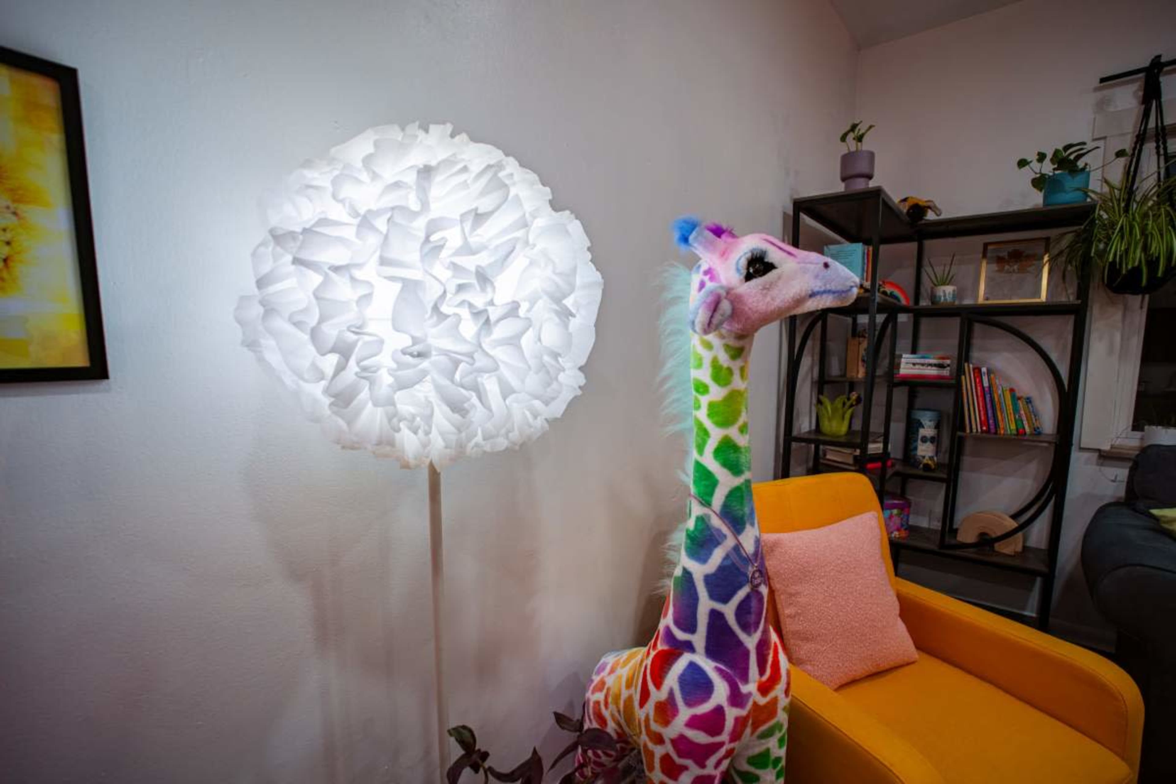 A colorful giraffe plush toy stands next to a round, white lamp in a cozy living room with a bookshelf and an orange armchair.