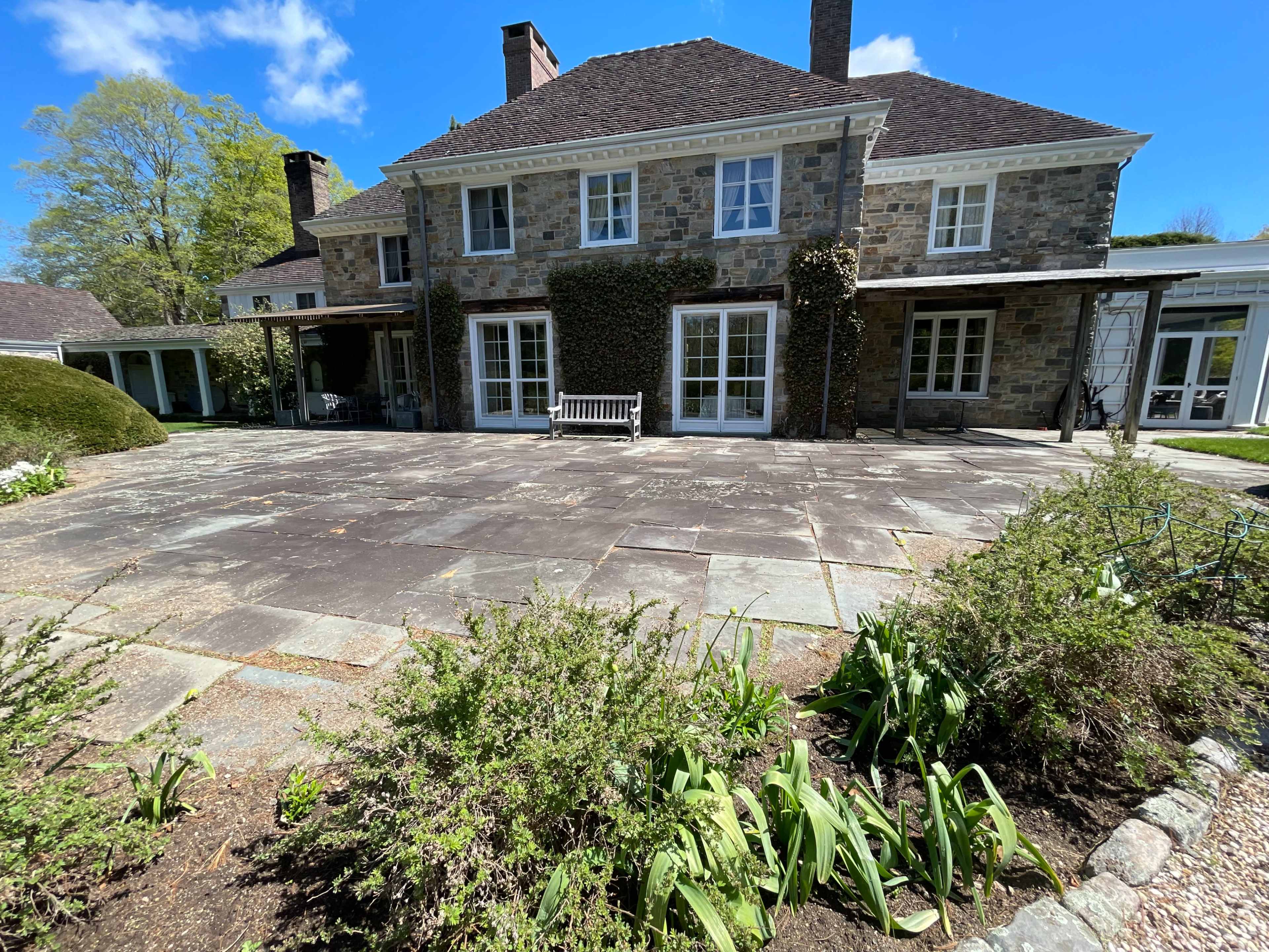 The image shows a stone house with multiple large windows, surrounded by a slate patio and landscaped garden areas.