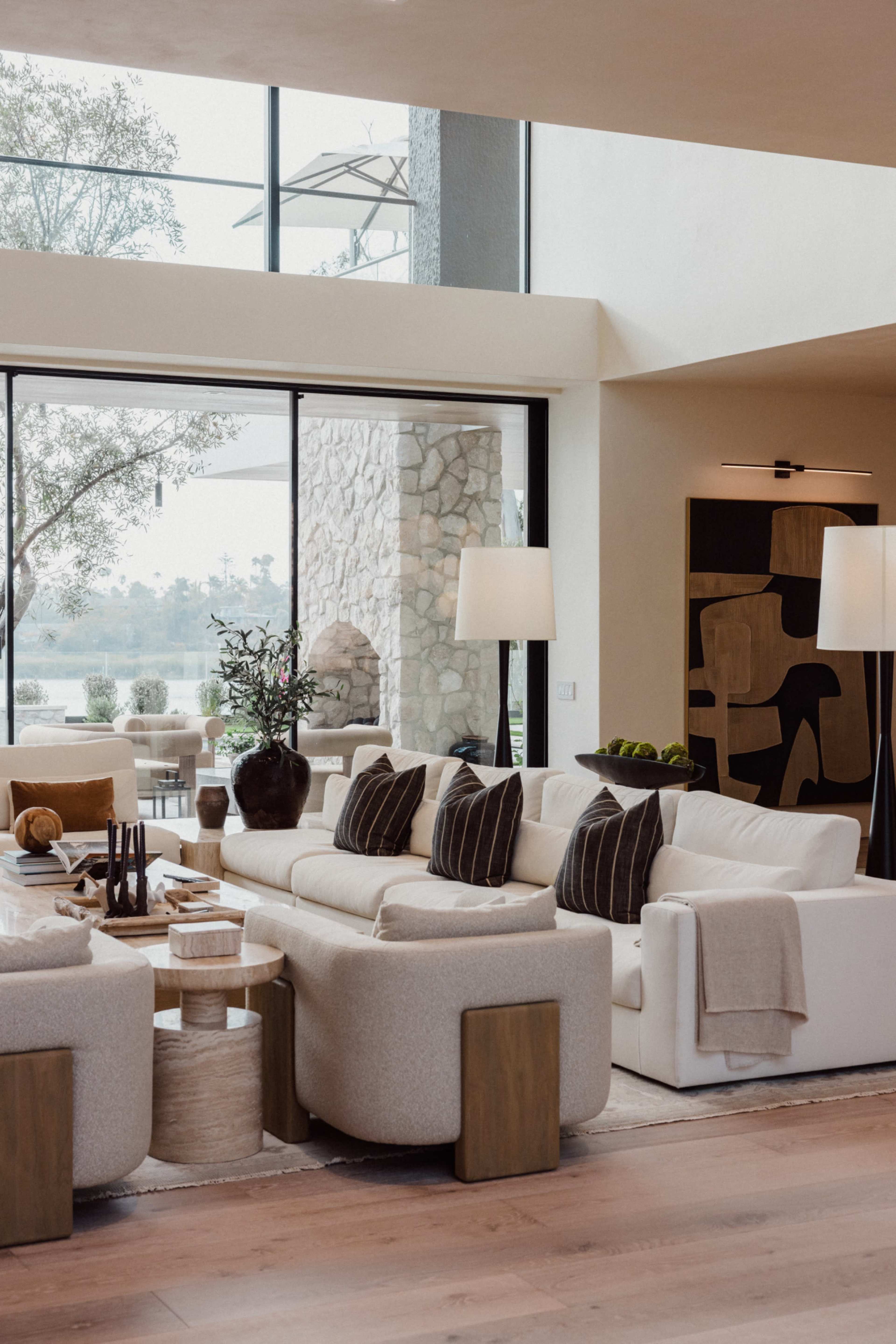 The image shows a modern living room with a white sofa, two light-colored chairs, and large windows that overlook a scenic outdoor area.