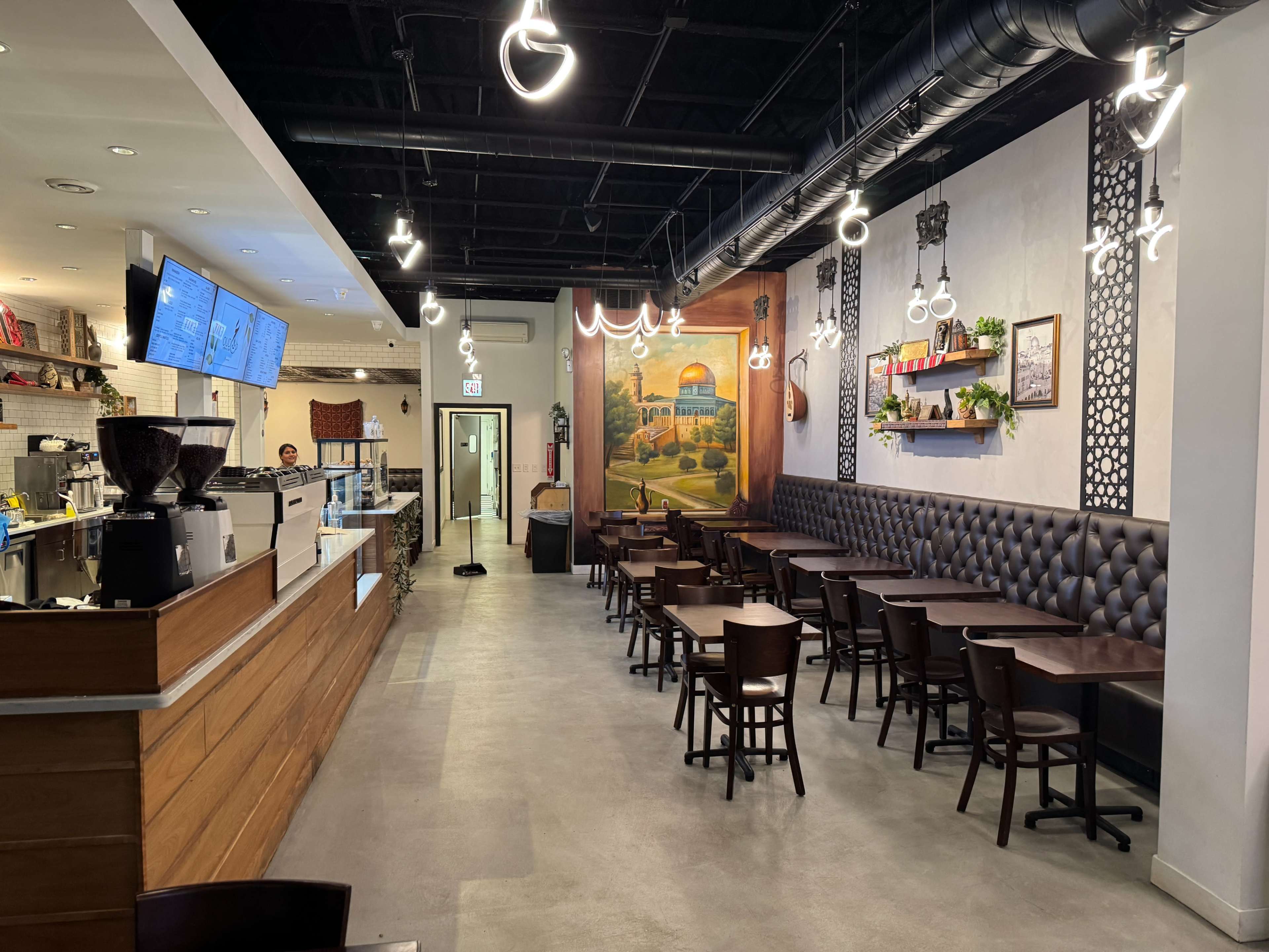 The image shows a modern café interior featuring wooden tables and chairs, a counter with a coffee machine, and decorative wall art.