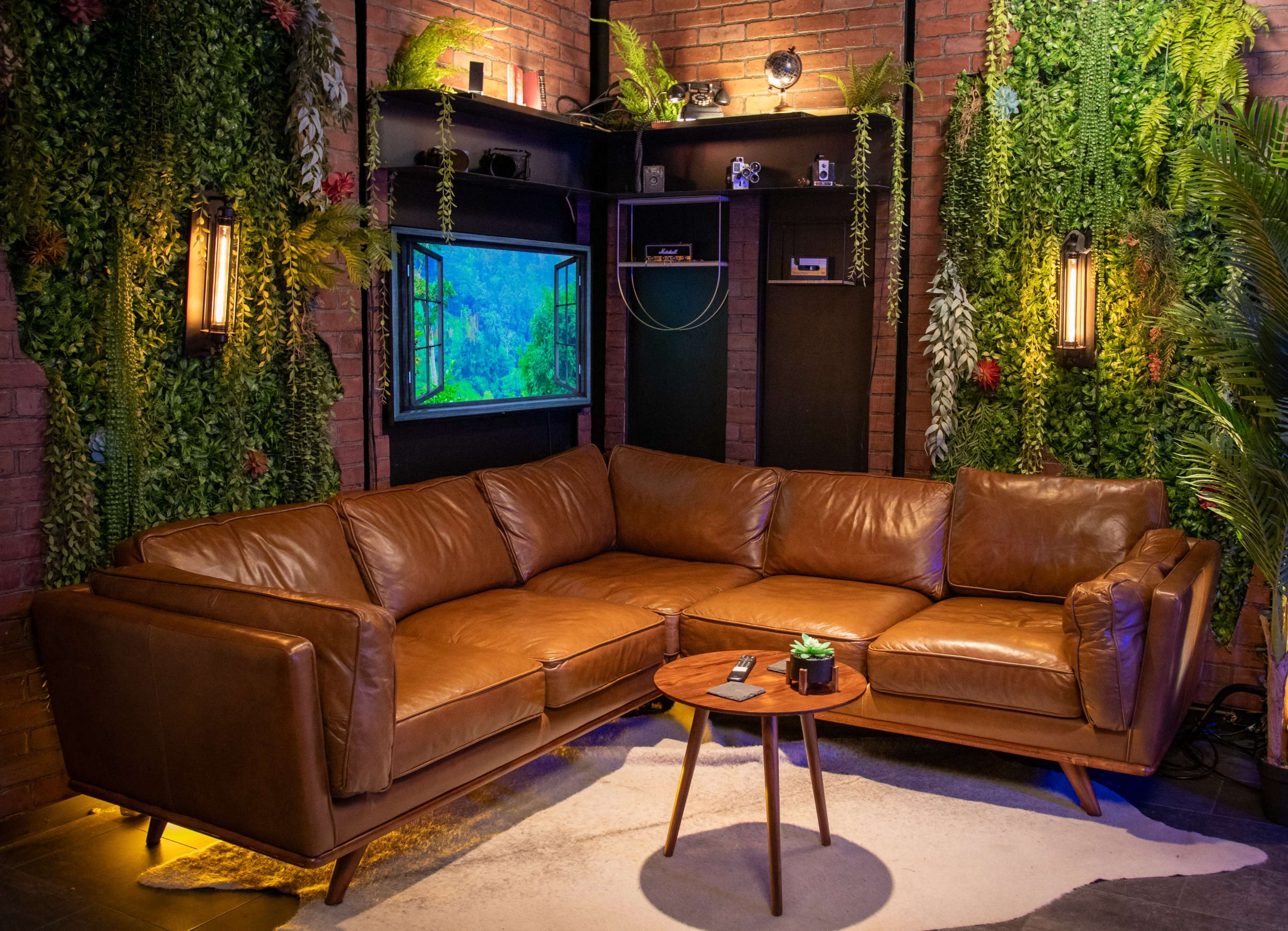 A cozy living room setup featuring a large brown leather sectional sofa, a small round wooden coffee table, and a flat-screen TV surrounded by green plants against a brick wall.