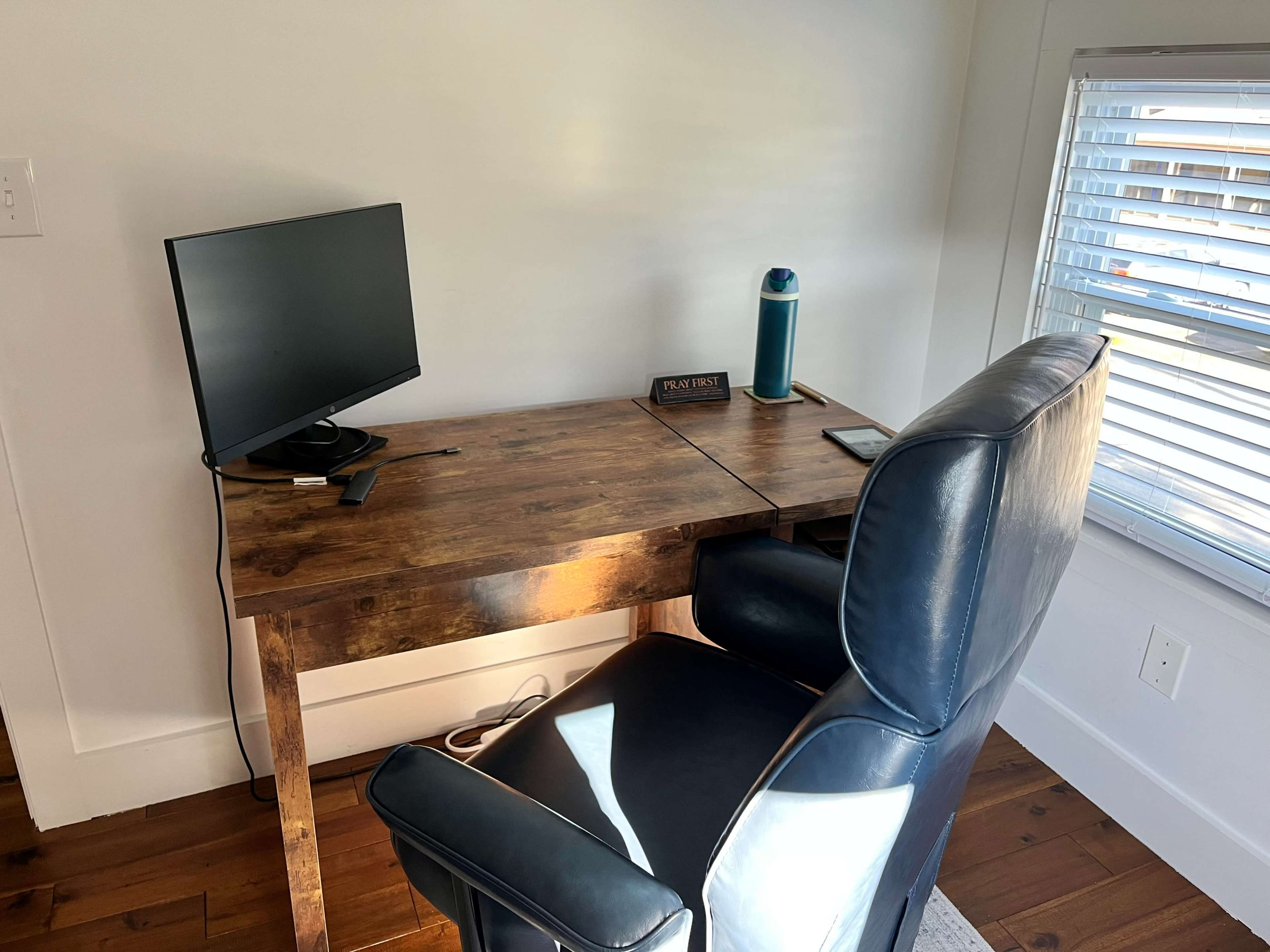 A wooden desk with a computer monitor, a water bottle, and a chair is positioned near a window with blinds.