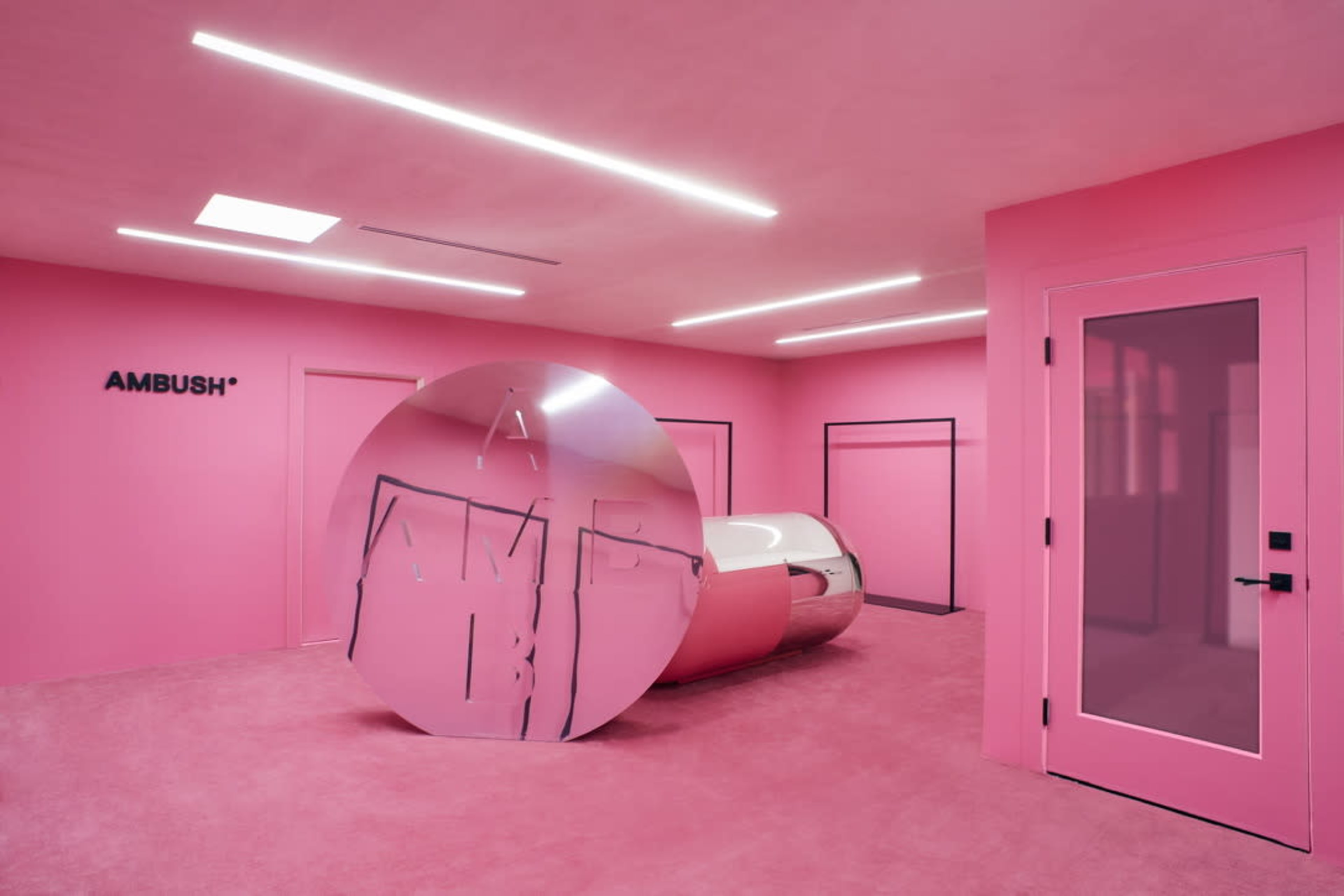 The image shows a modern, minimalist room with pink walls and flooring, featuring a large reflective sculpture and a sleek, round seating area.