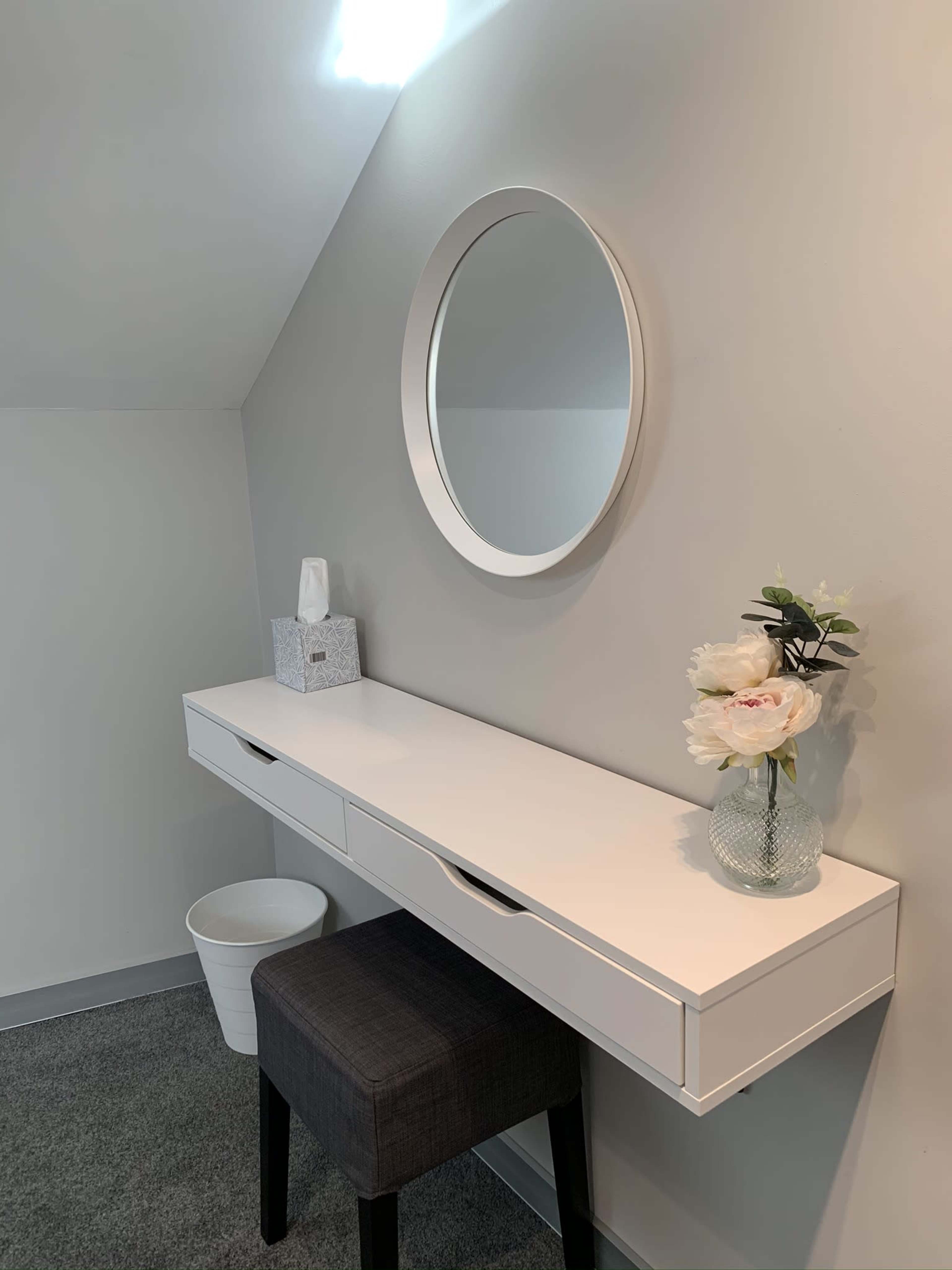 A minimalist vanity table with a round mirror, a vase of flowers, and a tissue box against a light gray wall.