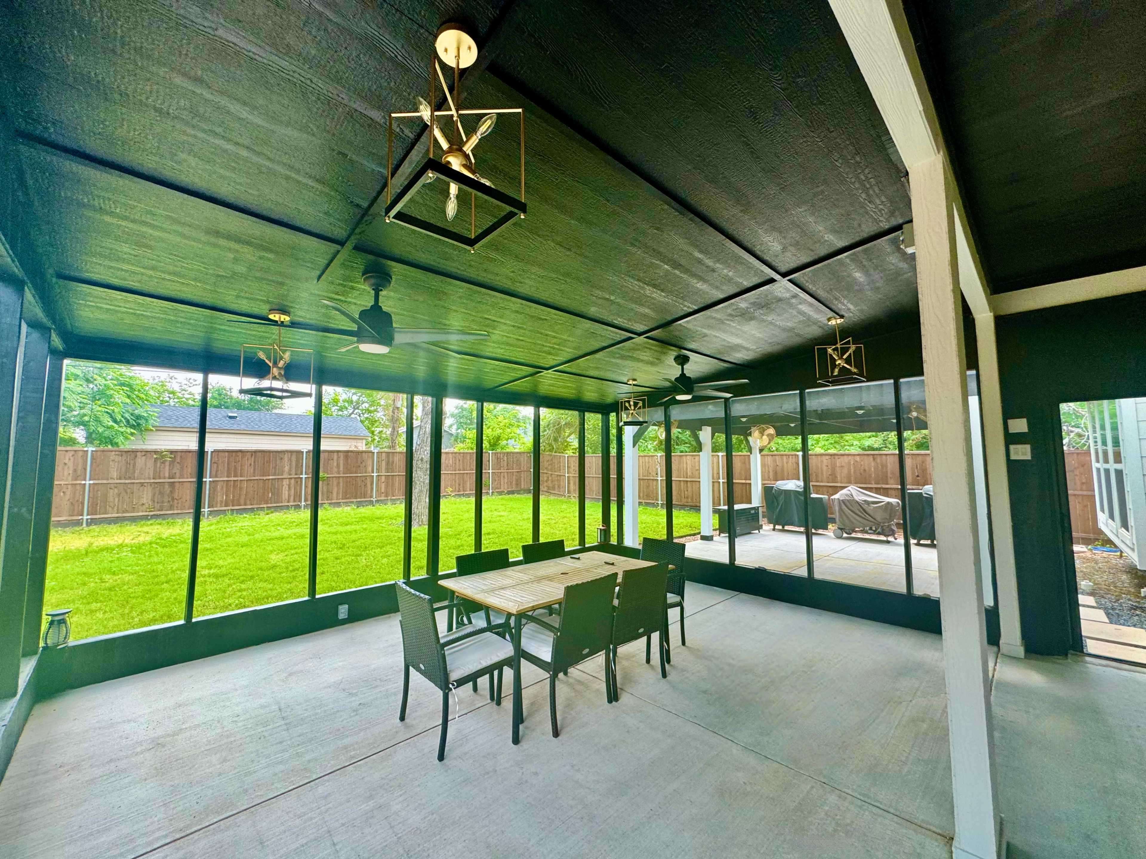 A screened-in patio with a table and chairs, ceiling fans, and a view of a backyard surrounded by a wooden fence.