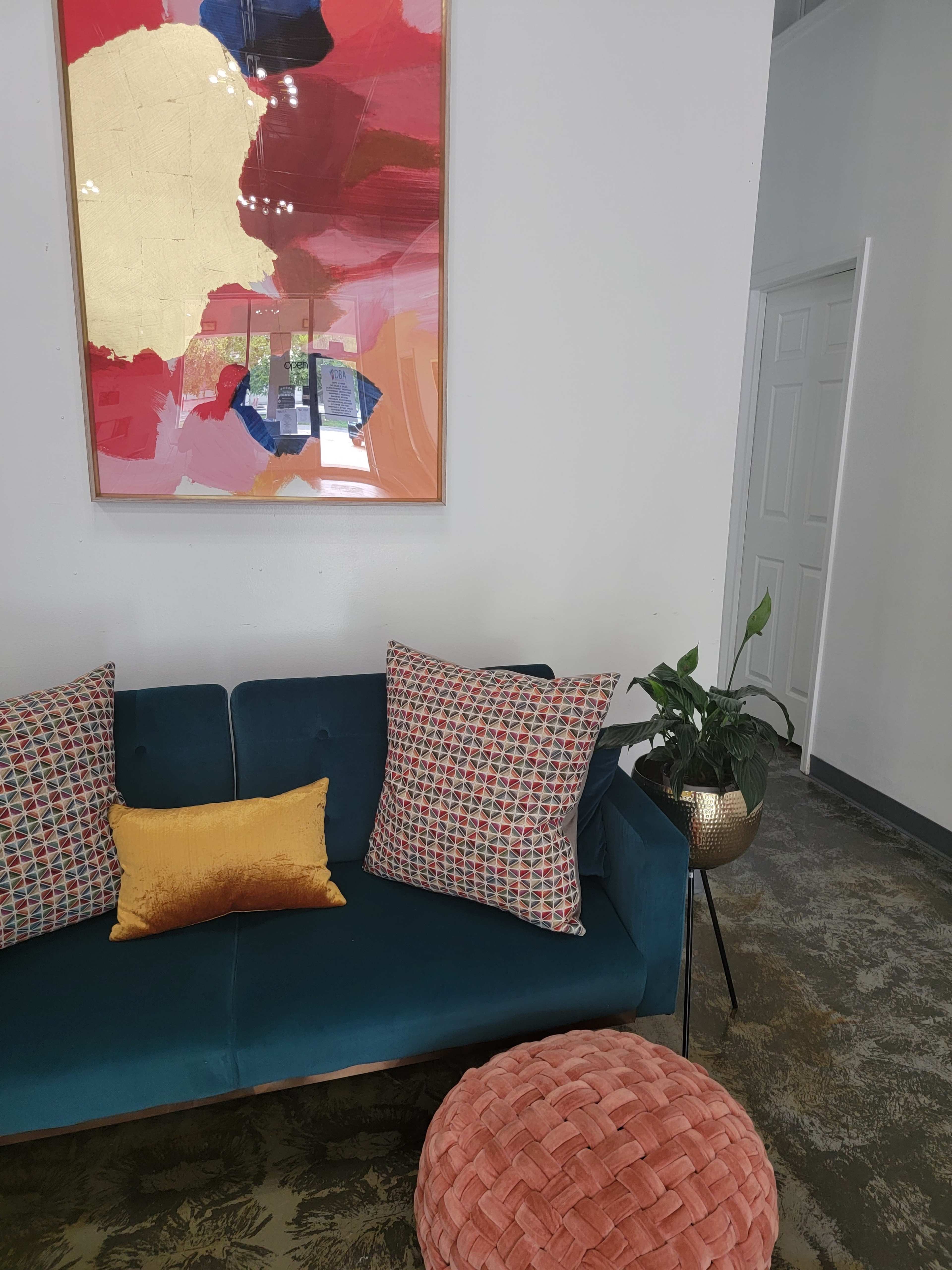 A teal sofa with patterned pillows and a yellow cushion is positioned in a room featuring a colorful abstract painting on the wall and a potted plant nearby.