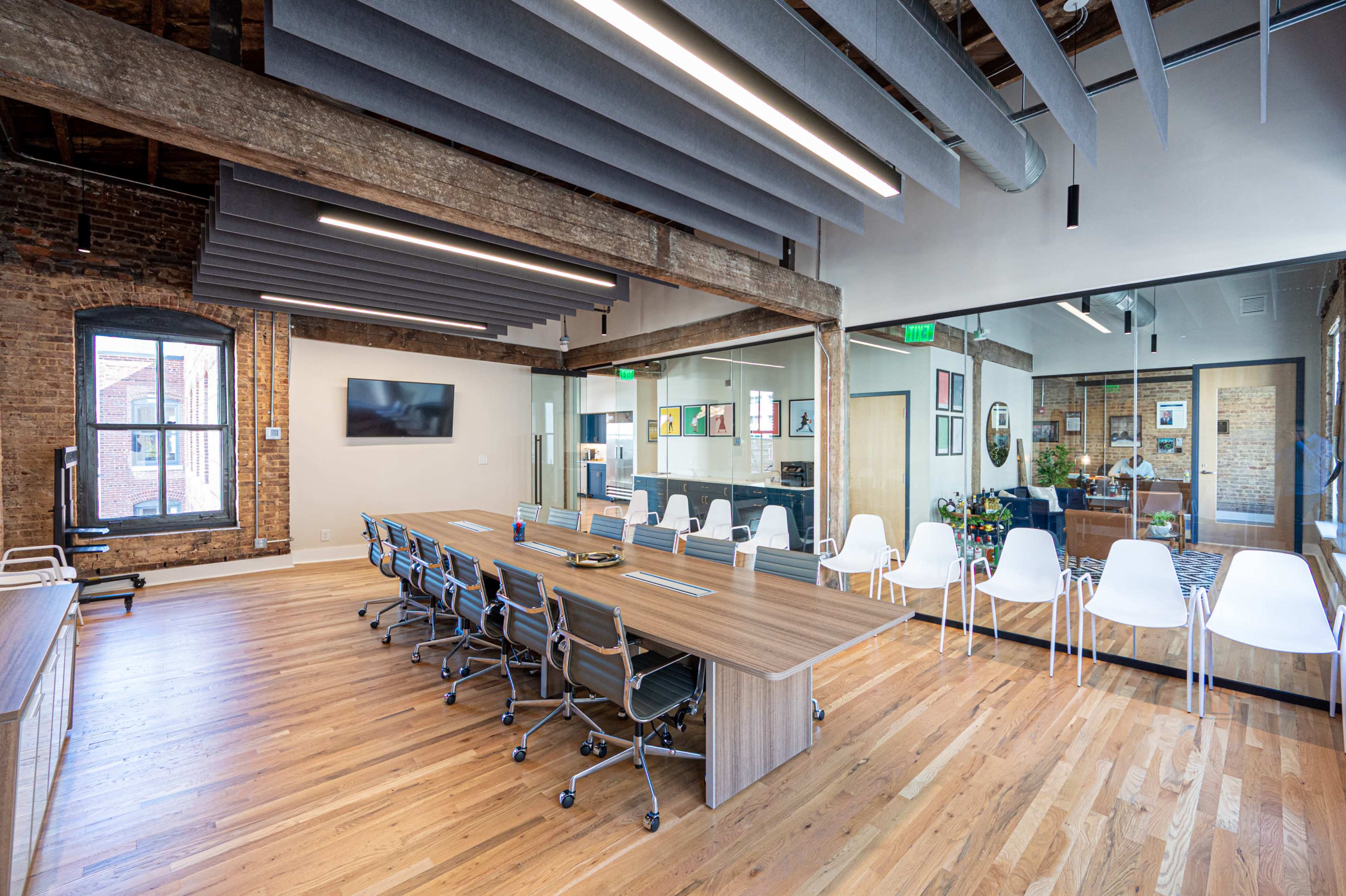 A modern conference room with a long wooden table surrounded by chairs, exposed brick walls, large windows, and a glass partition leading to another workspace.
