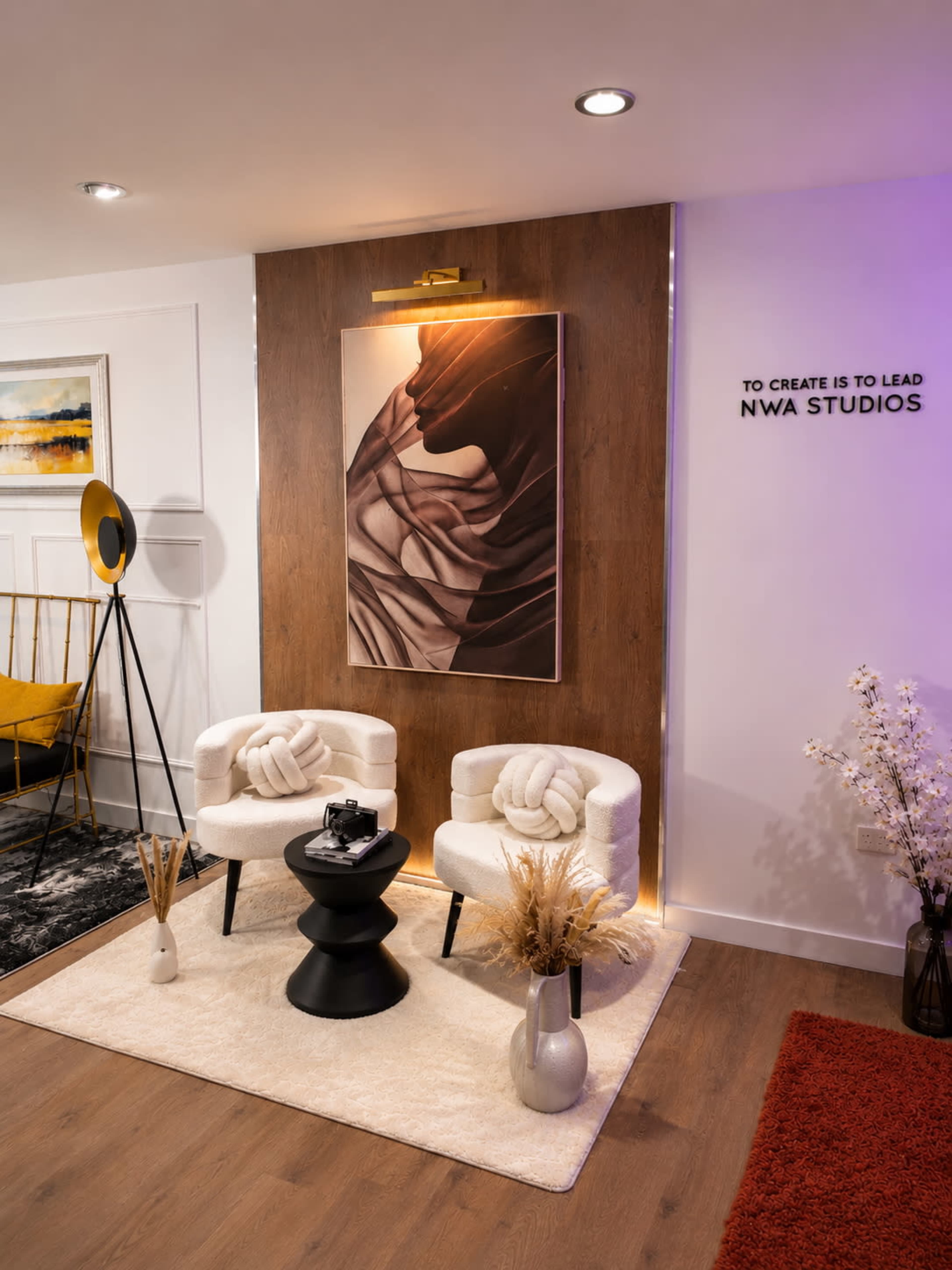 The image shows a modern, stylish studio space featuring two white chairs, a black table, an abstract artwork on the wall, and decorative elements including a floor lamp and plants.