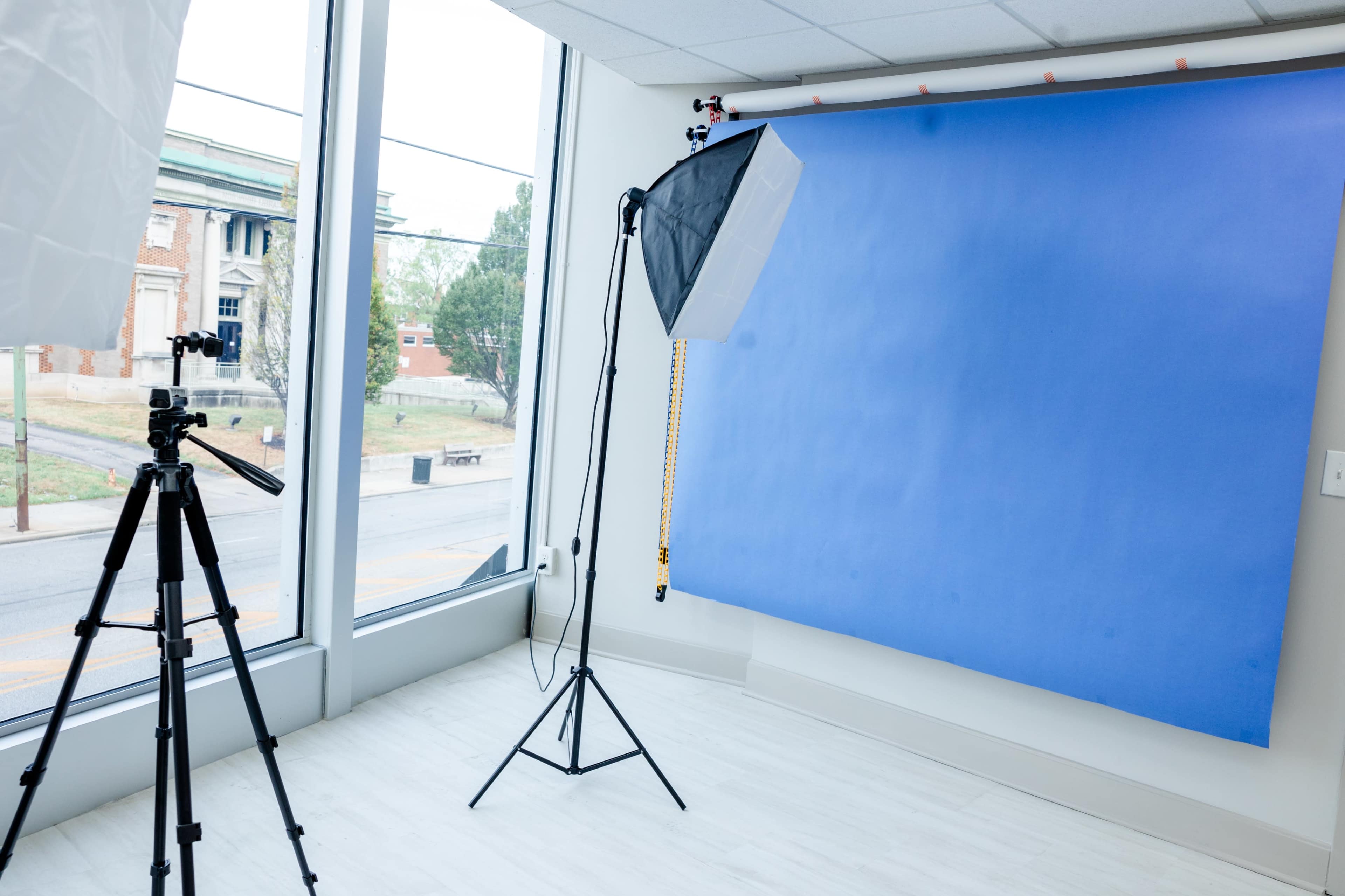 The image shows a photography studio setup with a blue backdrop, a lighting umbrella, and a camera on a tripod positioned beside a large window.