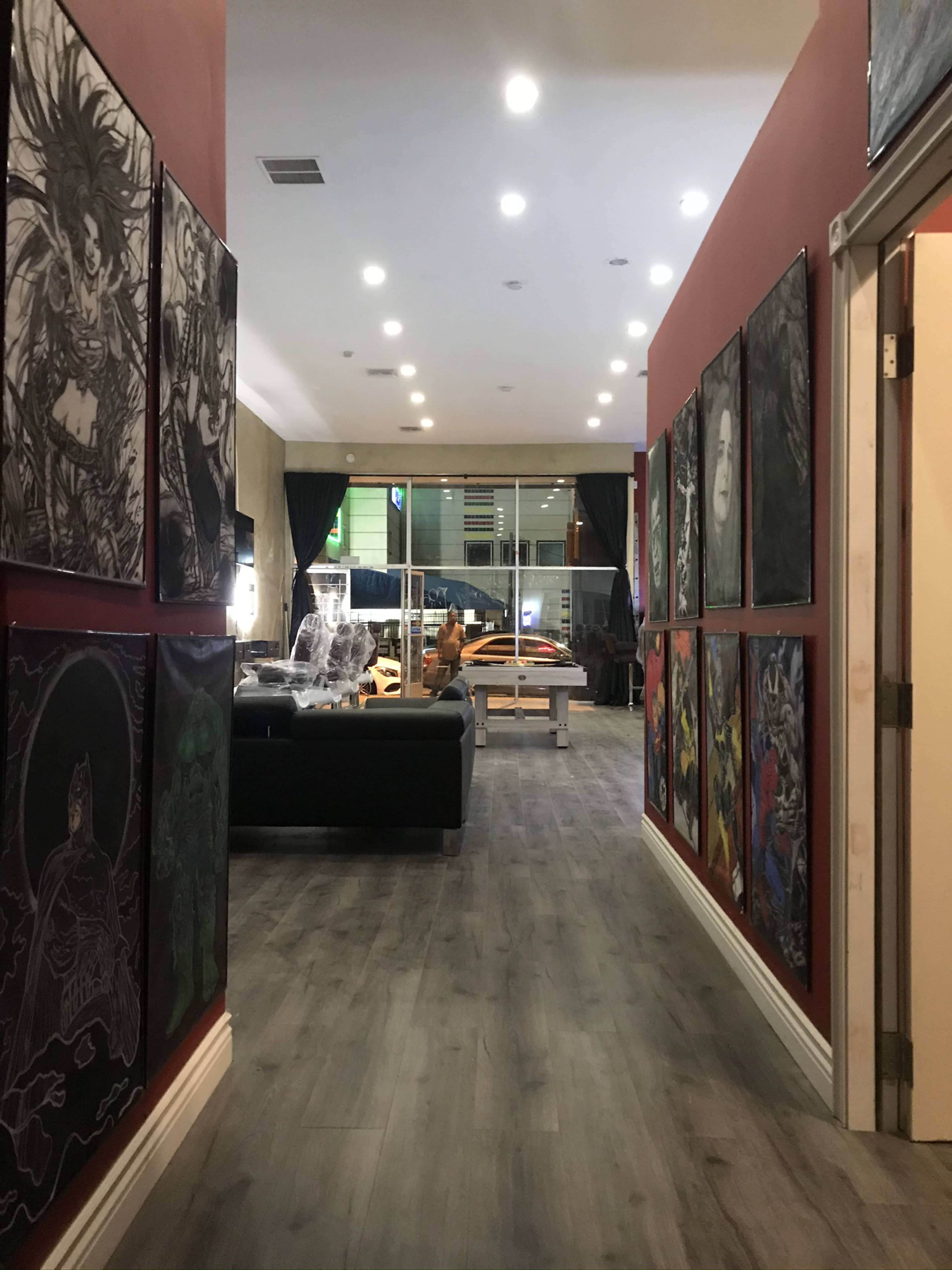 A hallway leading to a spacious room adorned with various framed artworks on the walls, alongside a black couch and large windows revealing vehicles outside.