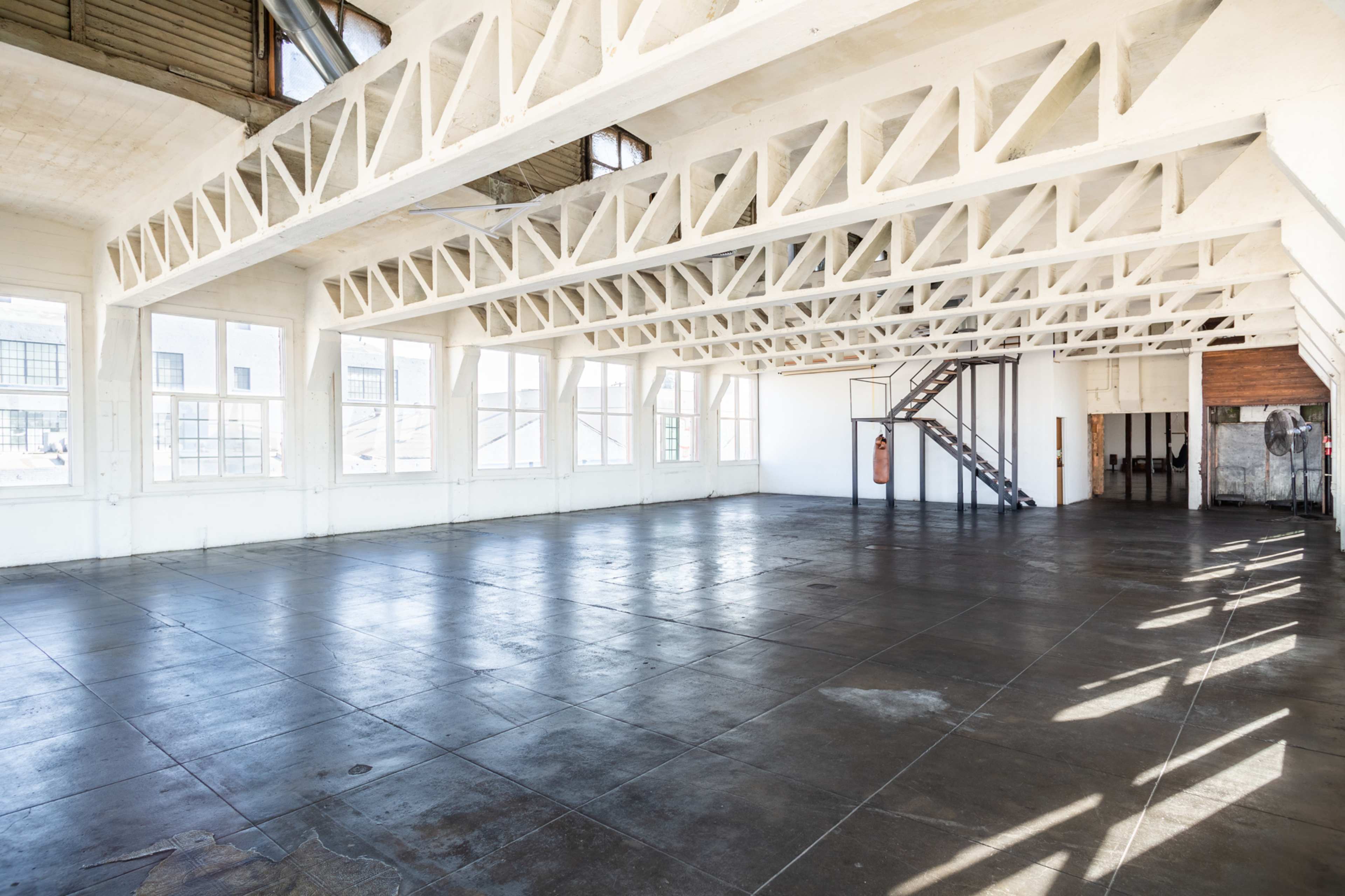 A spacious, empty industrial room with large windows, wooden beams, and a metal staircase.