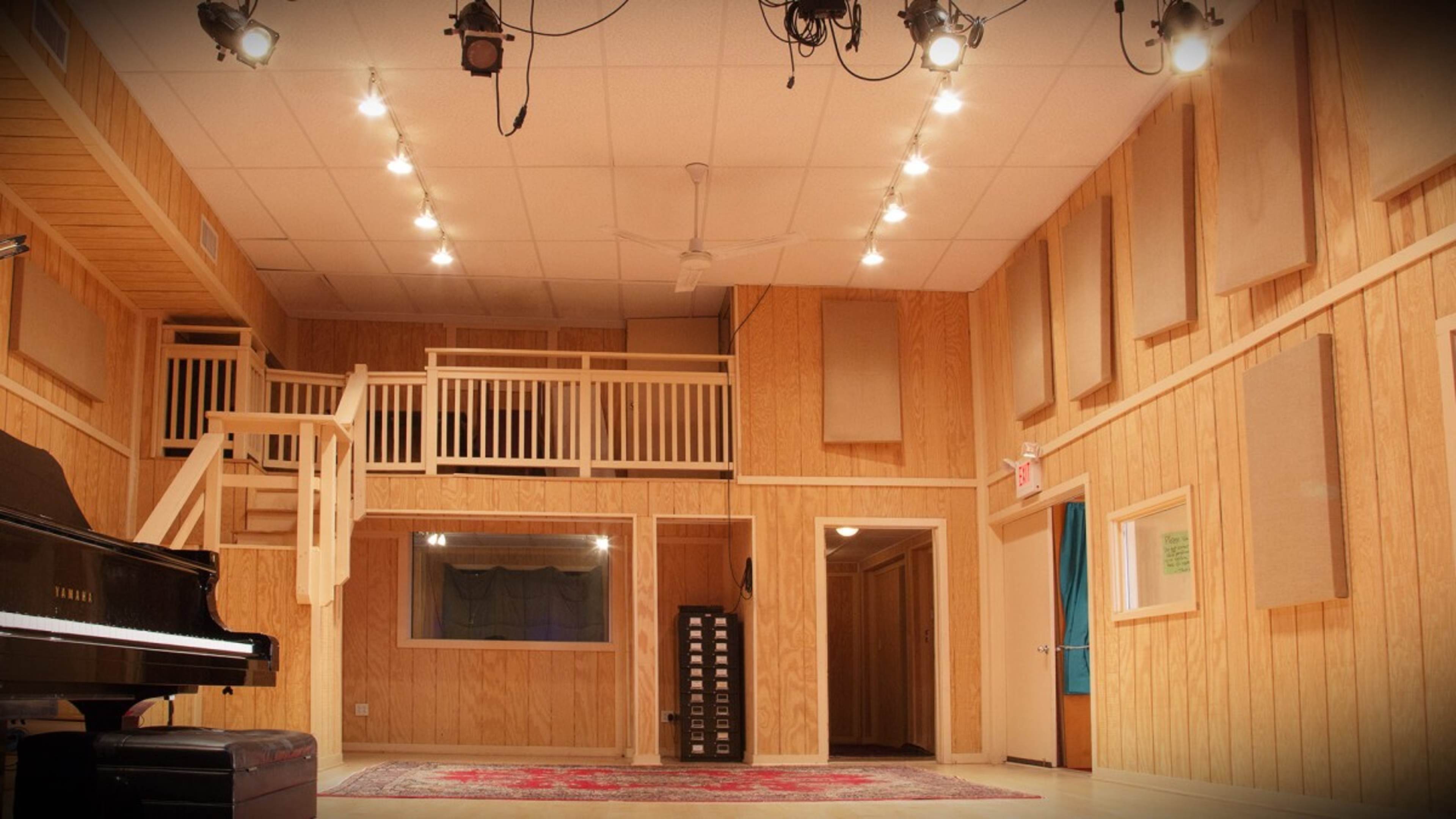 A wooden music studio with a grand piano in the foreground, a raised platform, and soundproofing panels on the walls.