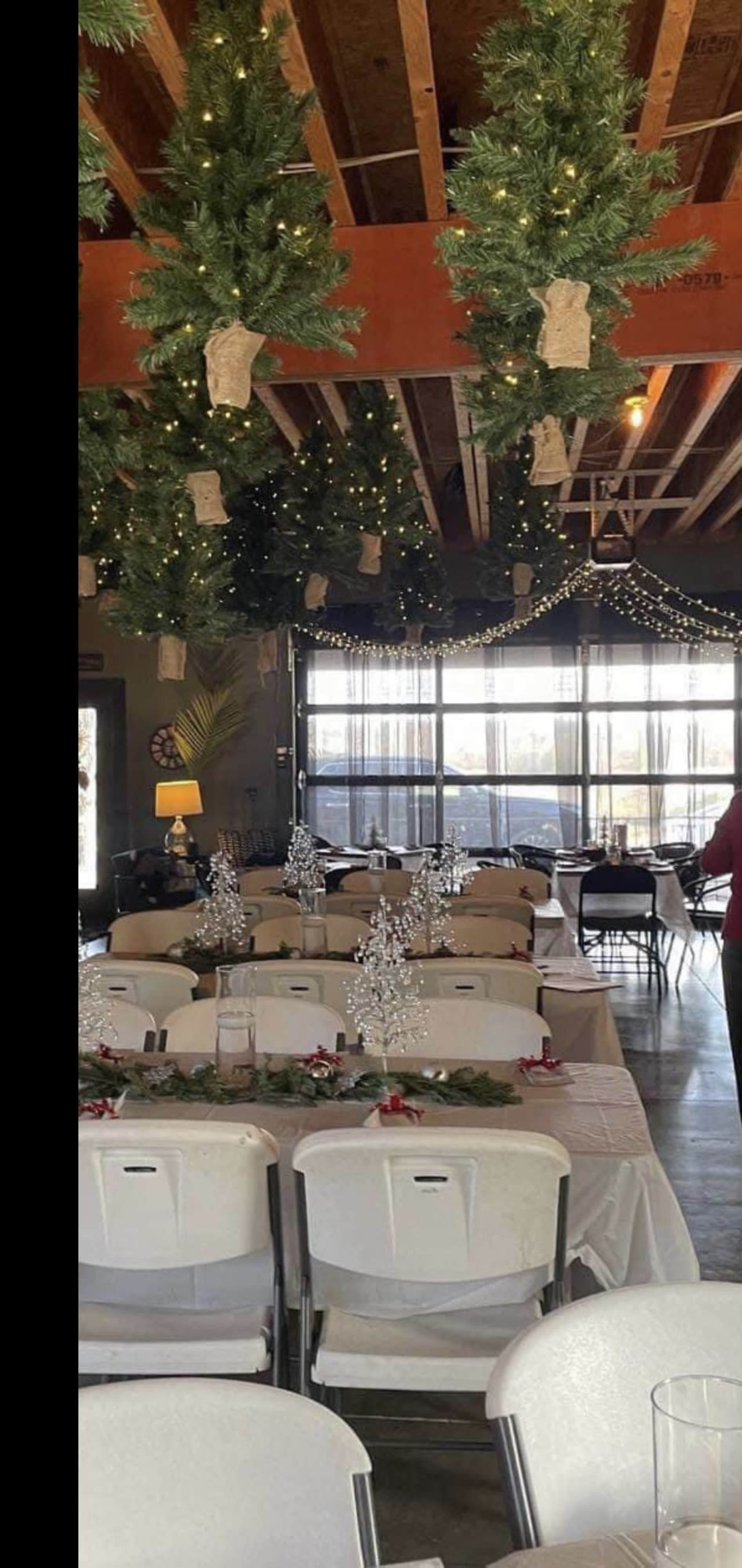 The image shows a decorated dining area with tables set for a gathering, featuring greenery and lights hanging from the ceiling.