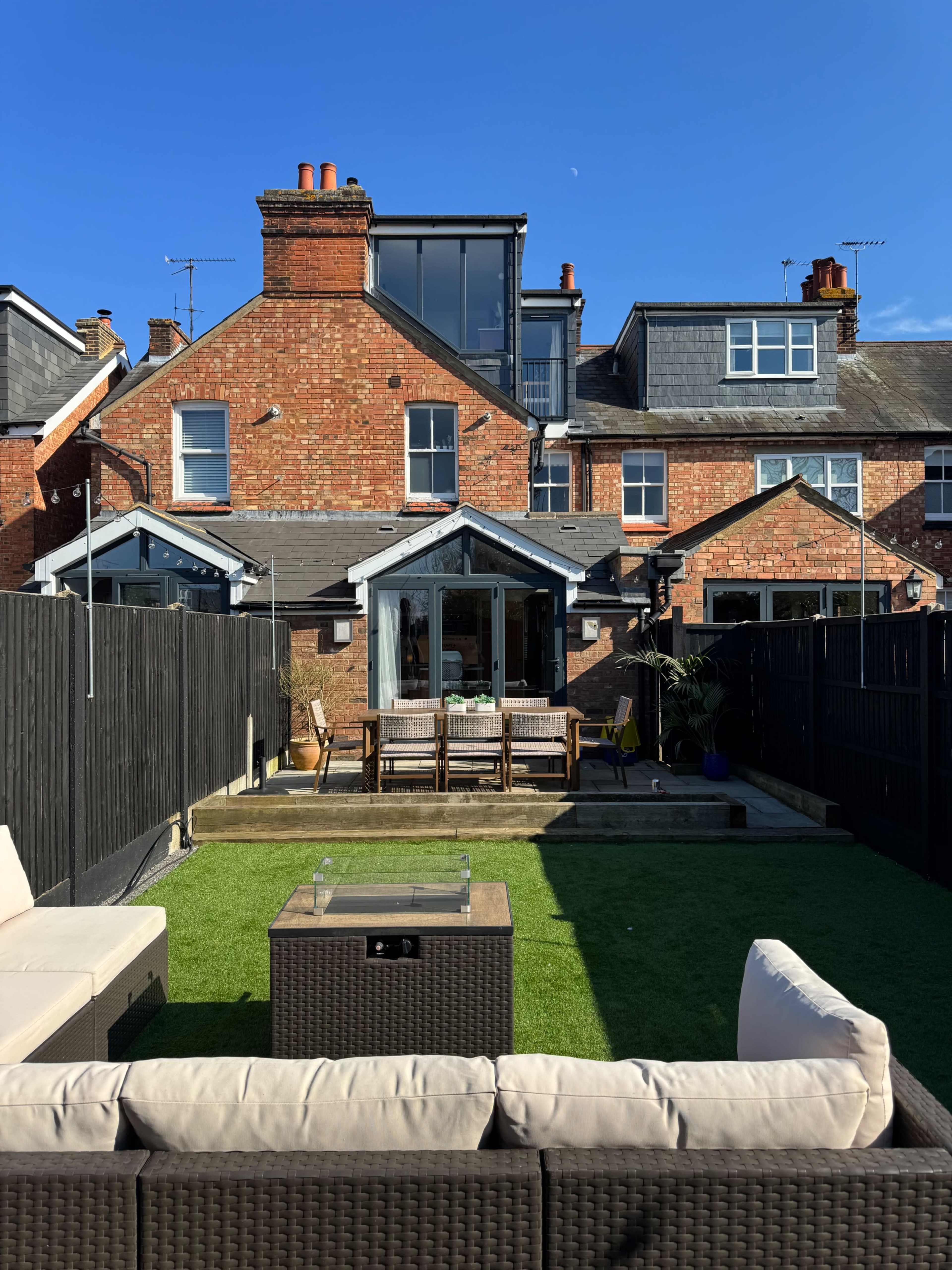 The image shows a backyard with a green lawn, seating area, and a brick house with multiple stories in the background.