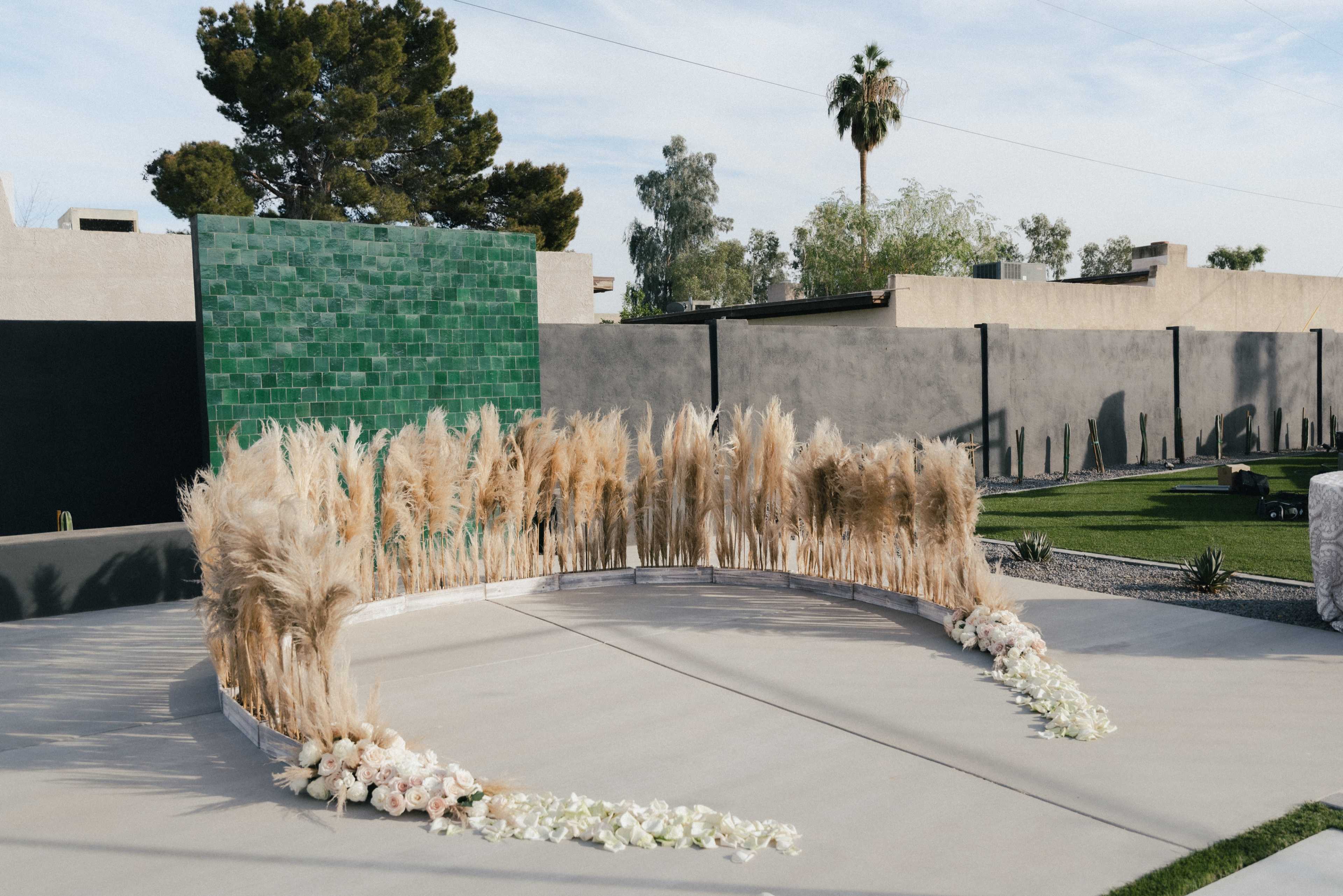 A circular outdoor ceremony setup featuring dried pampas grass, white floral arrangements, and a green tiled backdrop in a landscaped area.