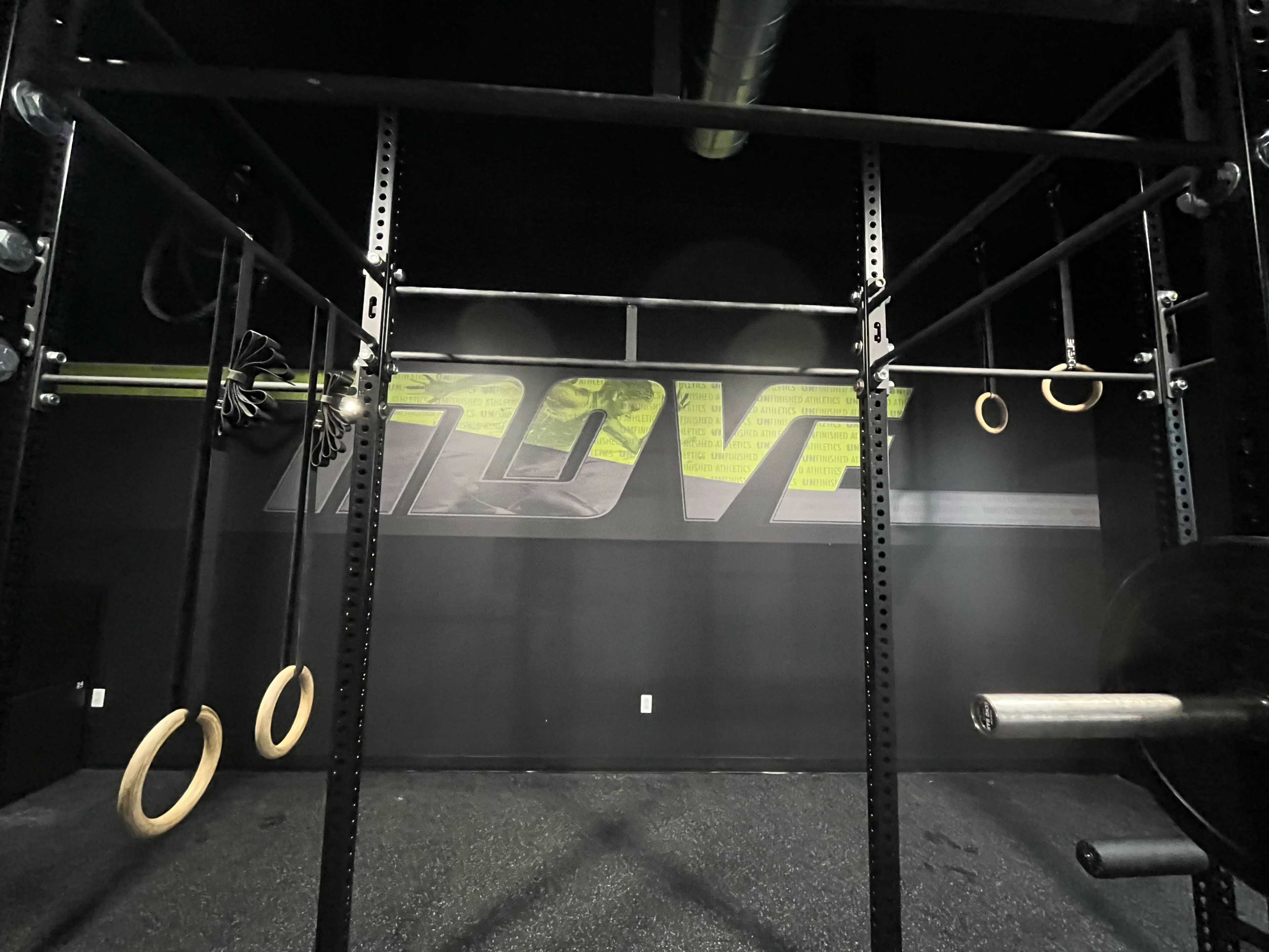 The image shows a gym space featuring a metal workout rig with hanging gymnastic rings and a wall displaying the word "MOVE."