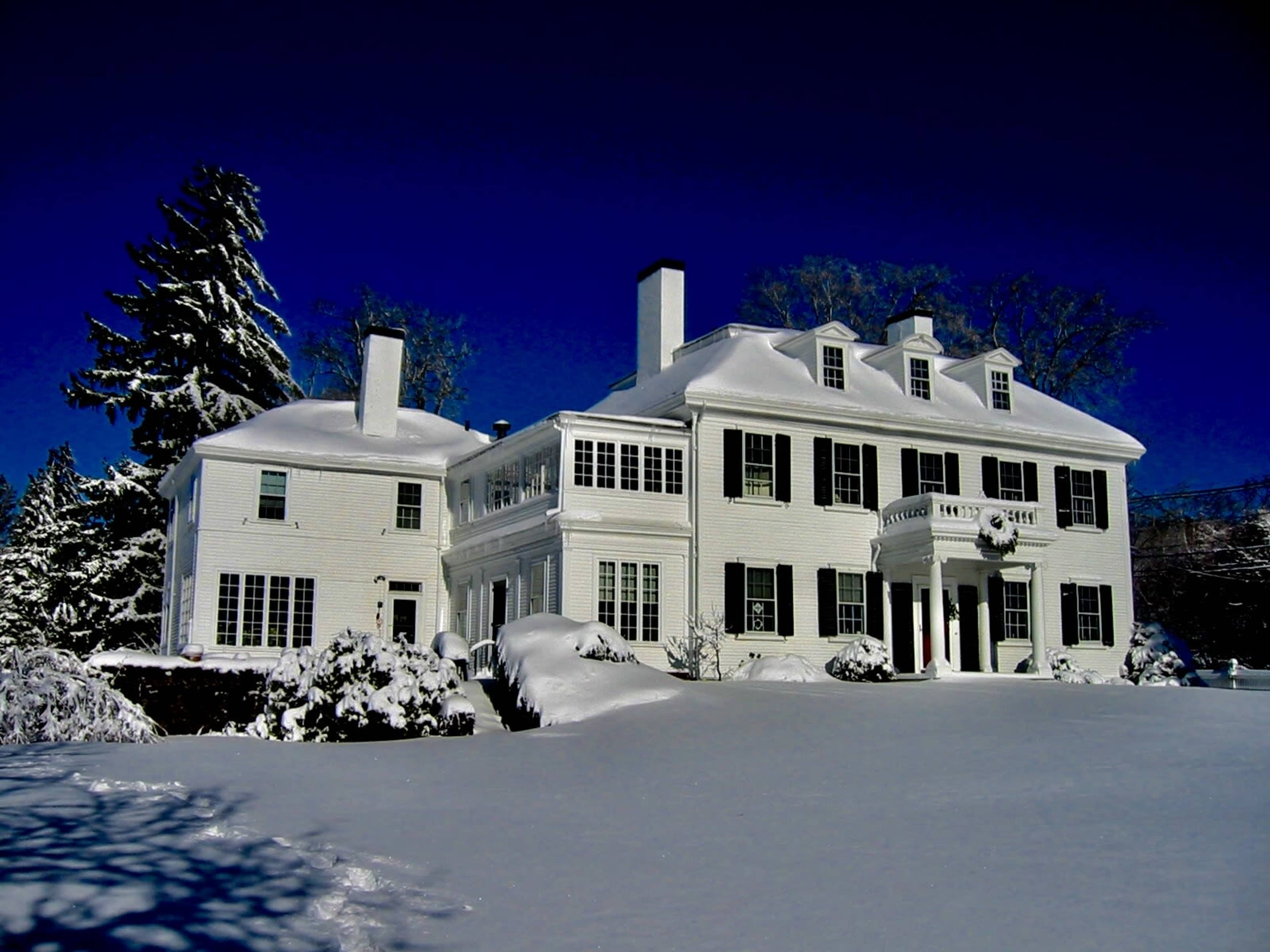 A large white colonial-style house is surrounded by deep snow, with tall pine trees in the background under a clear blue sky.