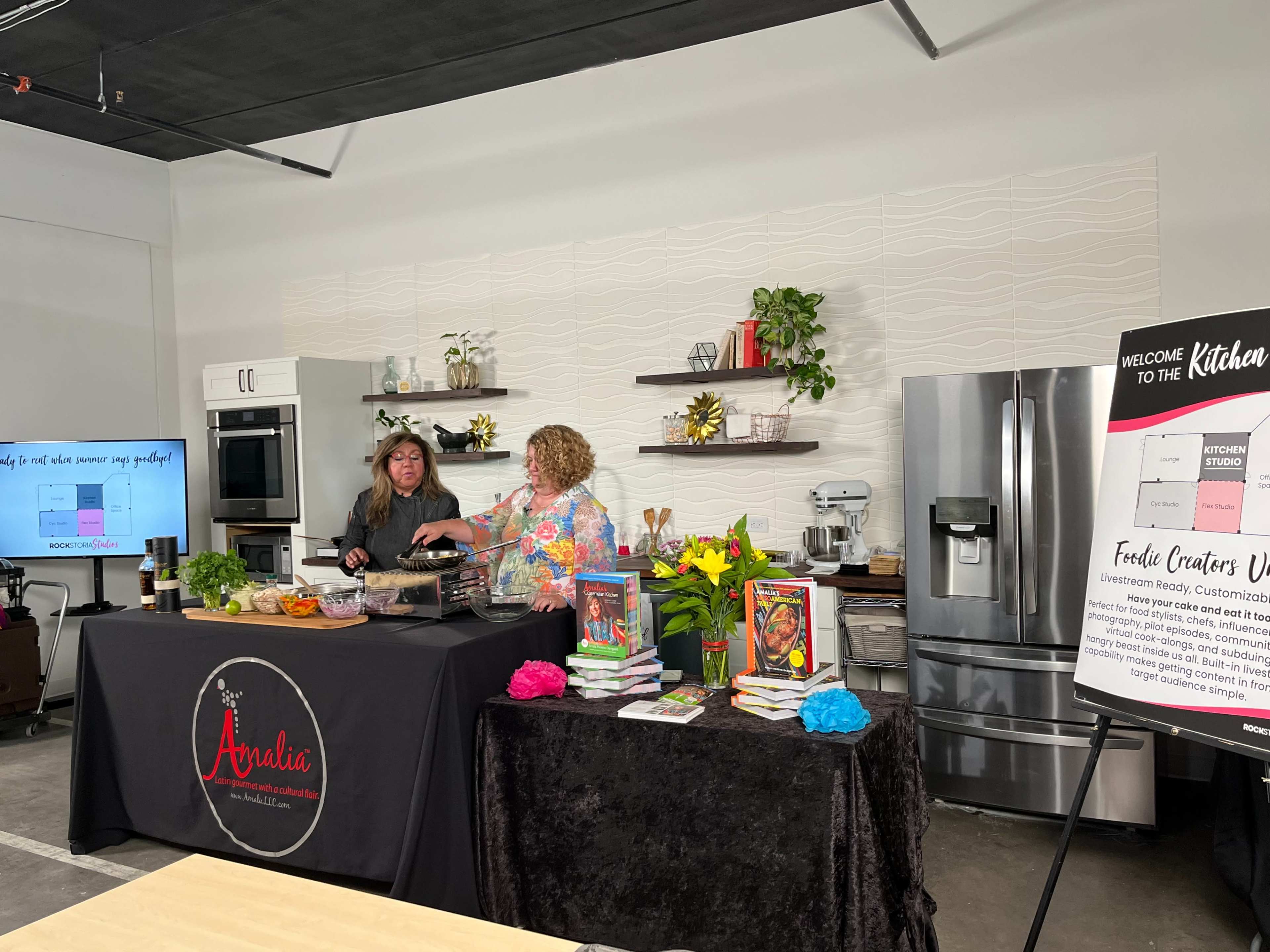Two women are preparing food at a kitchen counter decorated with plants and cookbooks, while a large sign welcomes visitors to the cooking space.