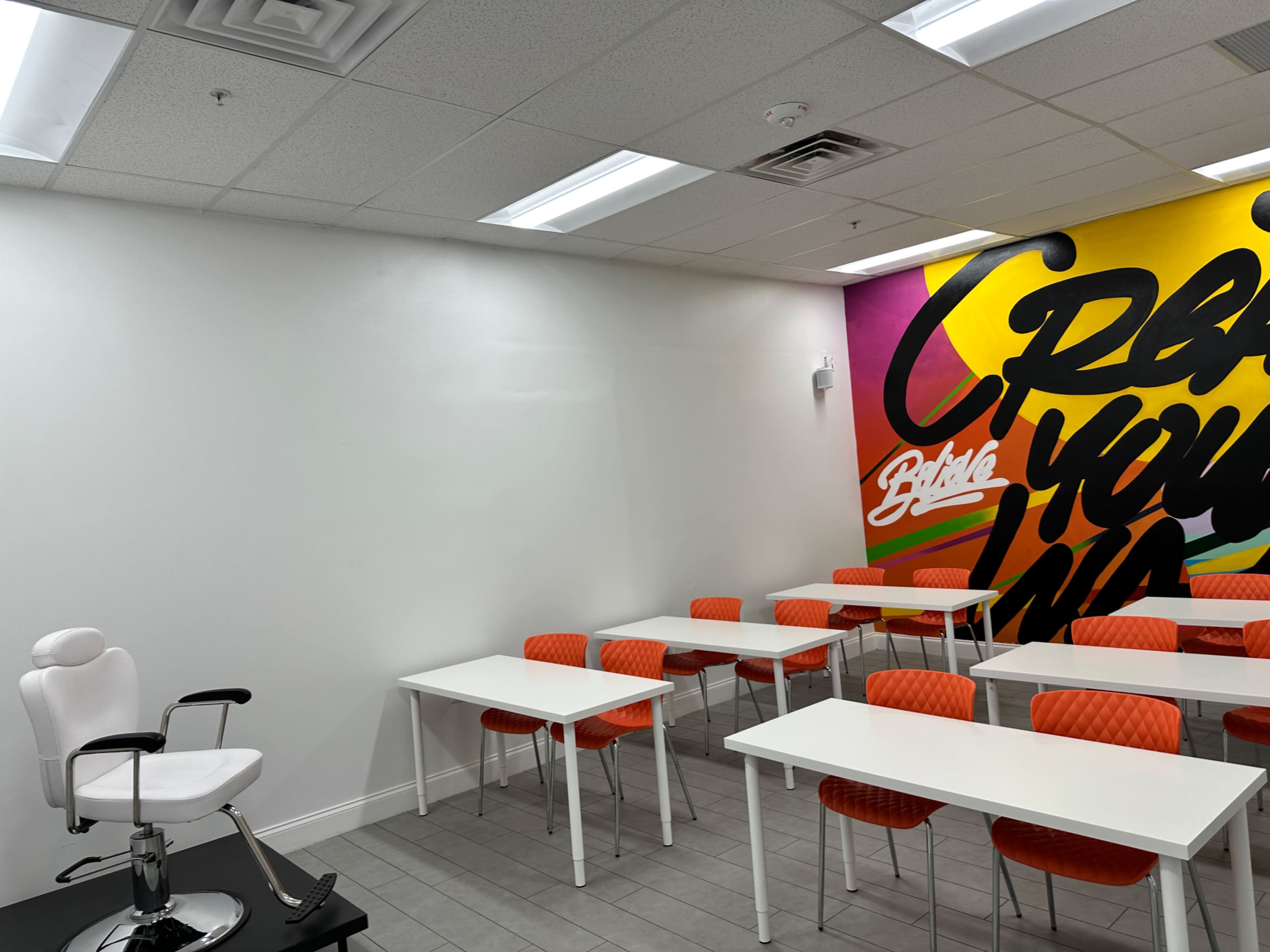 A brightly colored classroom with a white styling chair on one side and rows of white tables with orange chairs.