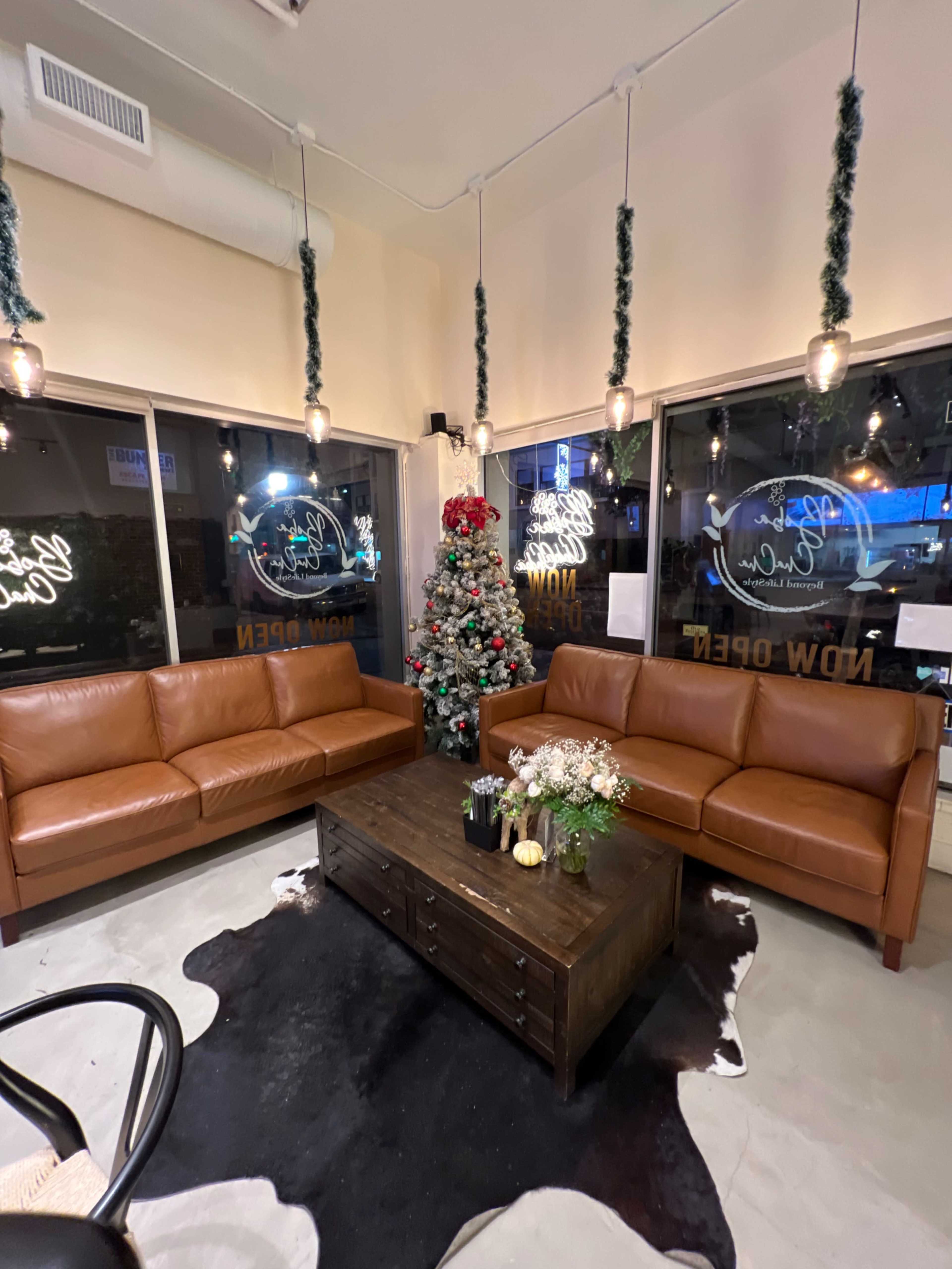 A cozy seating area with two brown leather sofas facing a wooden coffee table, a decorated Christmas tree, and large windows featuring neon signs.
