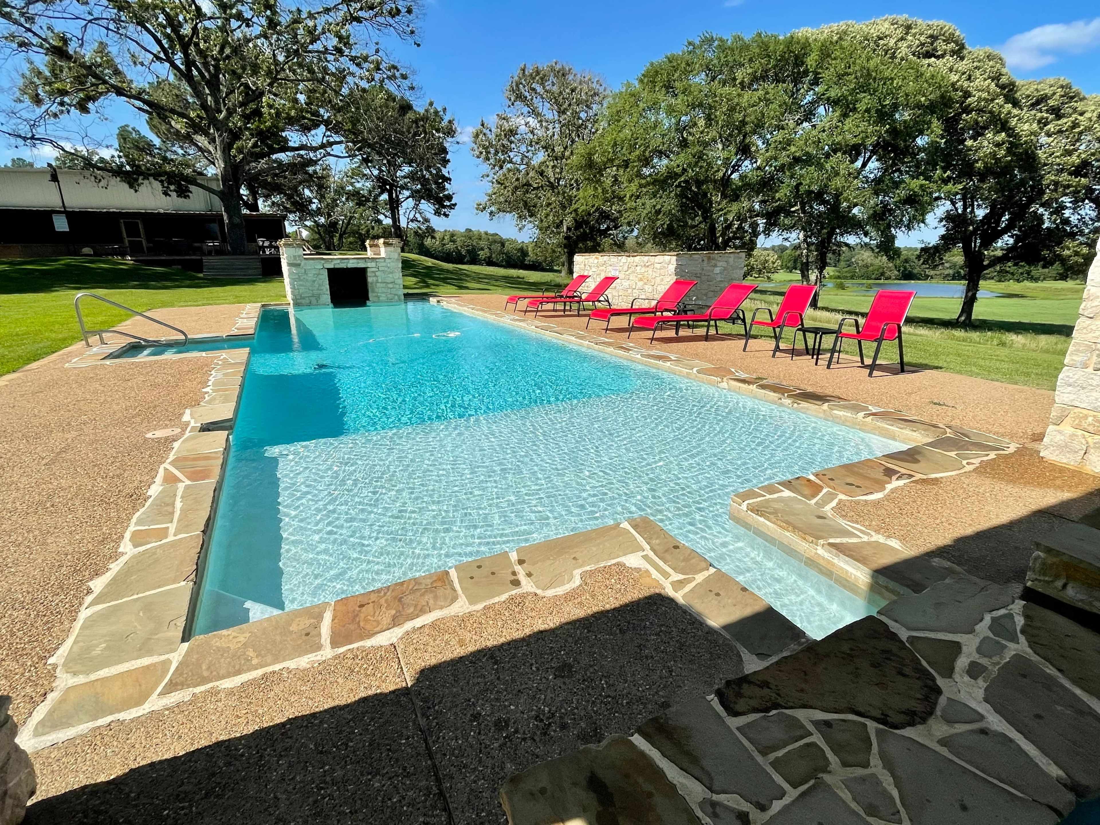 The image shows a clear swimming pool surrounded by stone patio, lounge chairs, and greenery.