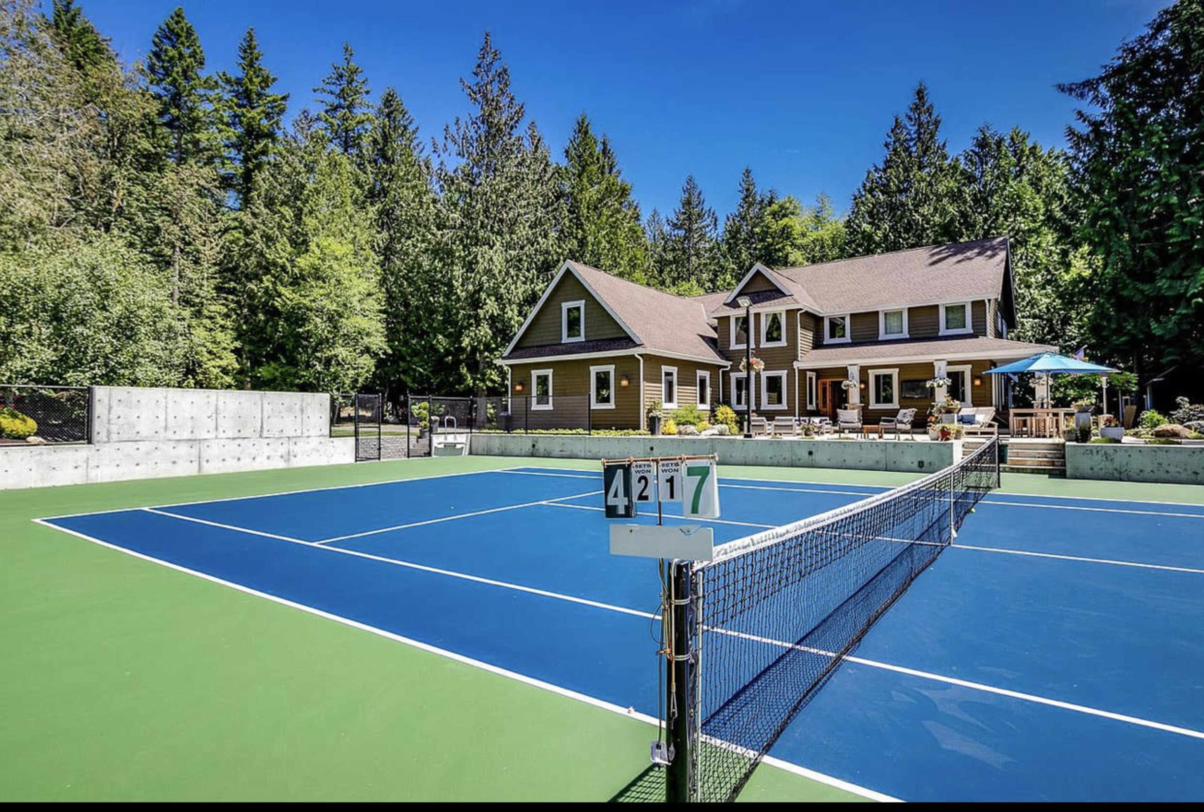 The image shows a well-maintained tennis court in front of a large, two-story house surrounded by tall trees.