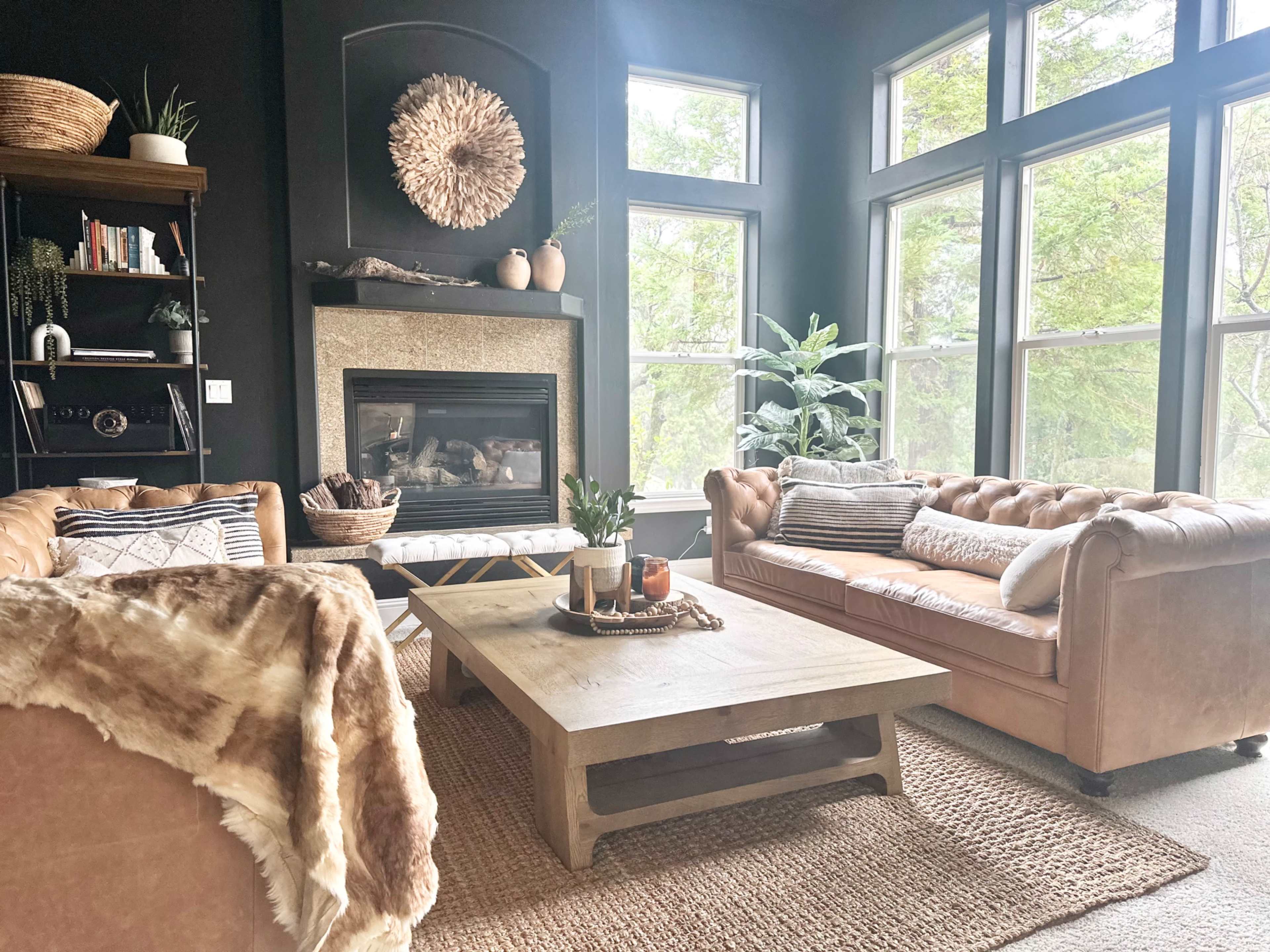 A cozy living room features a stone fireplace surrounded by large windows, two leather sofas, a wooden coffee table, and decorative plants.