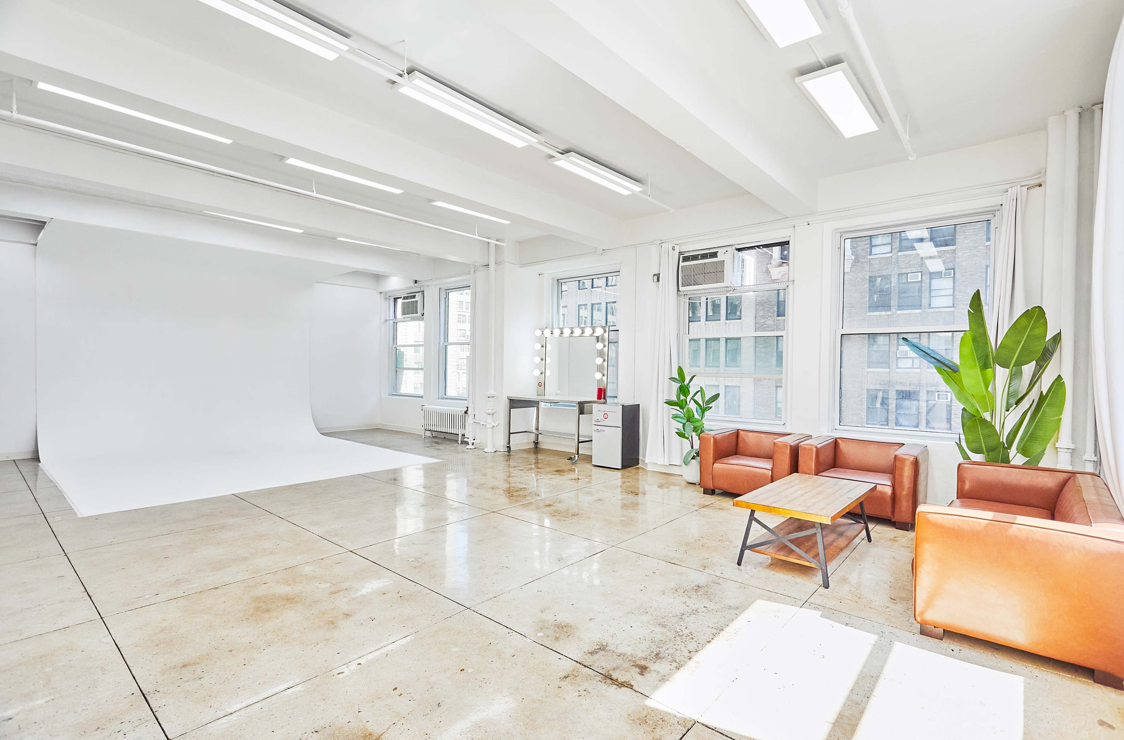 The image shows a bright, spacious photography studio featuring a white backdrop, a seating area with brown leather couches, a coffee table, and large windows.