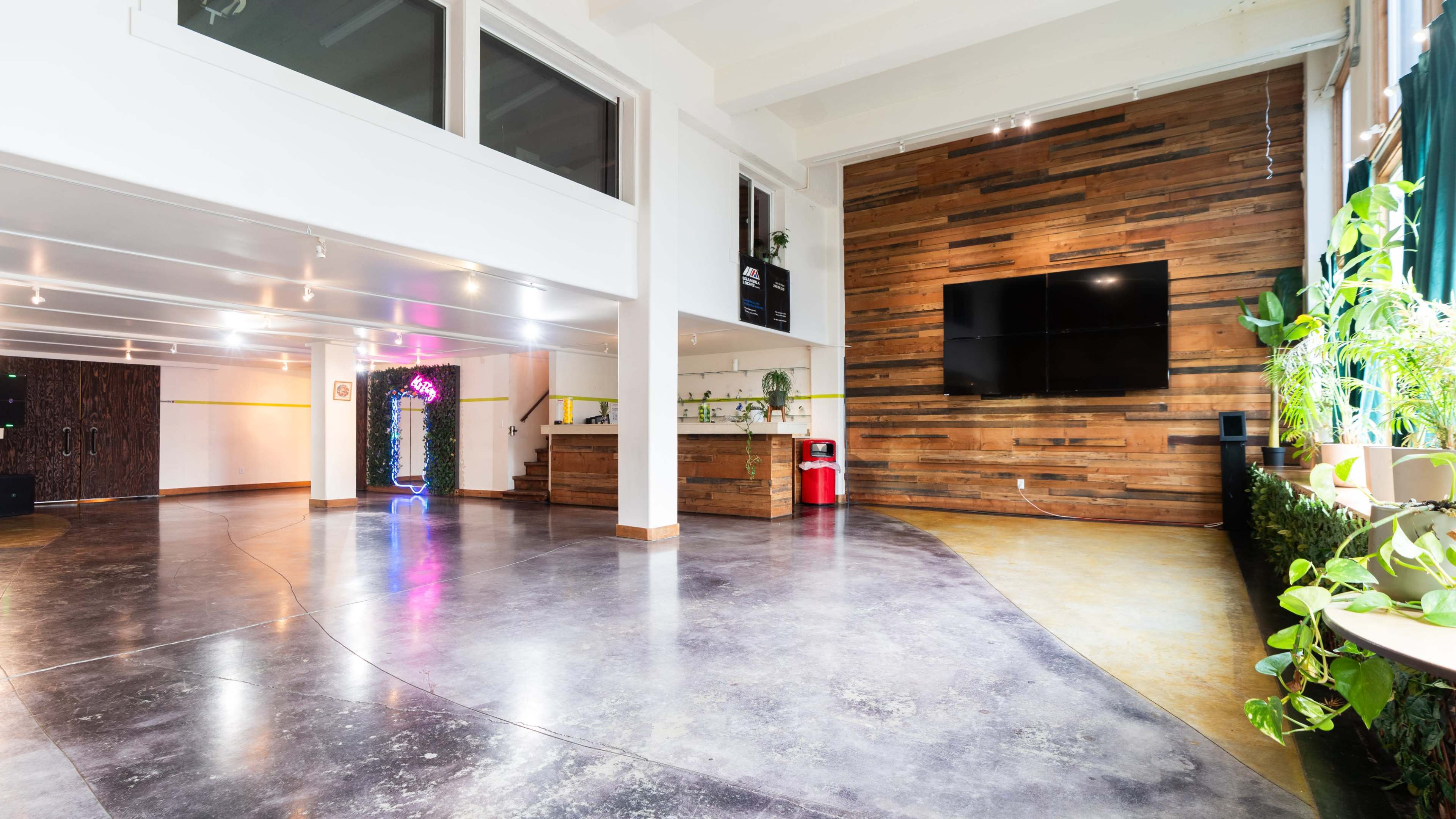 The image shows a modern interior space with polished concrete floors, a wooden accent wall, a large screen mounted on the wall, and various plants.