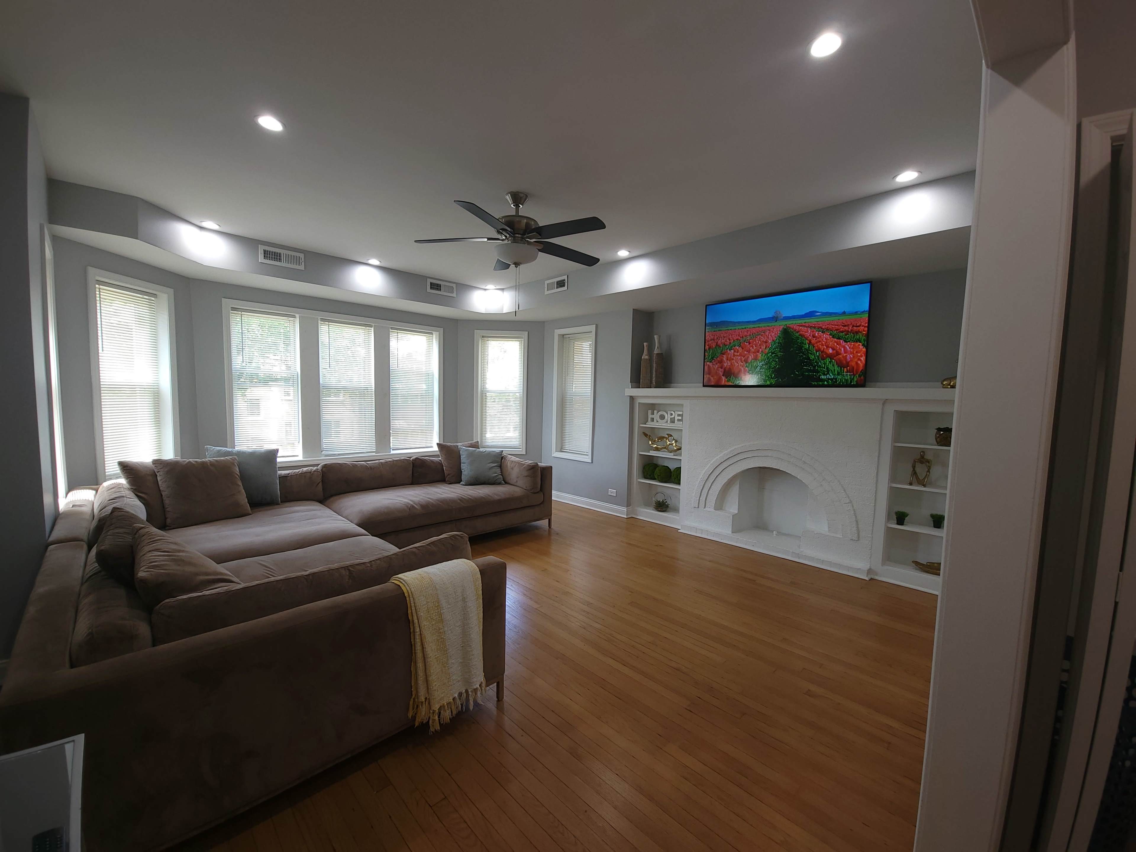 The living room features a large sectional sofa, a white fireplace with built-in shelves, and a wall-mounted television above the fireplace, illuminated by recessed lighting.