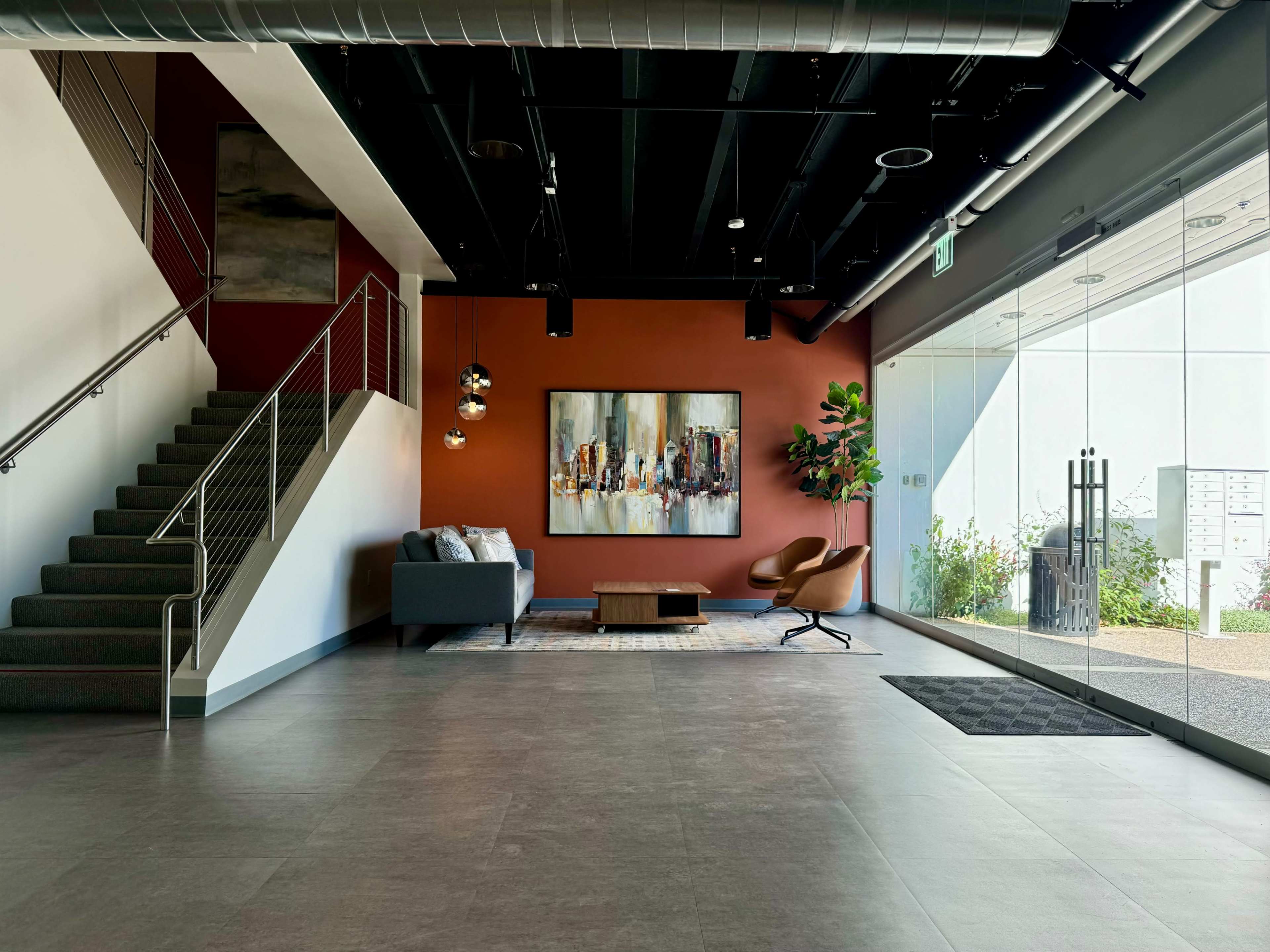 The image shows a modern lobby area featuring a staircase, a couch, an armchair, a coffee table, and a large abstract painting on the wall.