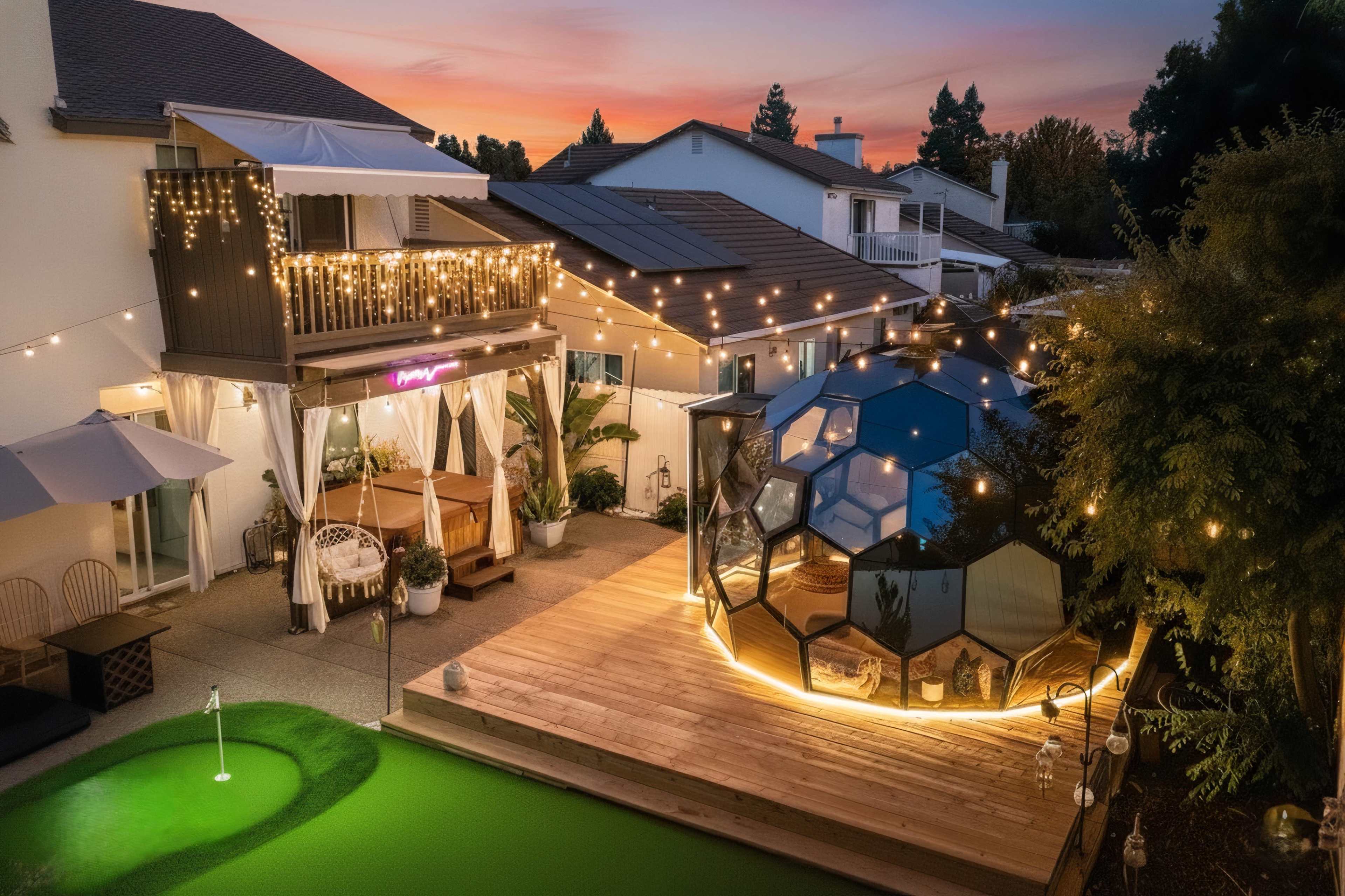 The image shows a backyard scene with a modern, hexagon-shaped structure, a raised wooden deck, and outdoor lighting, all set against a colorful sunset sky.