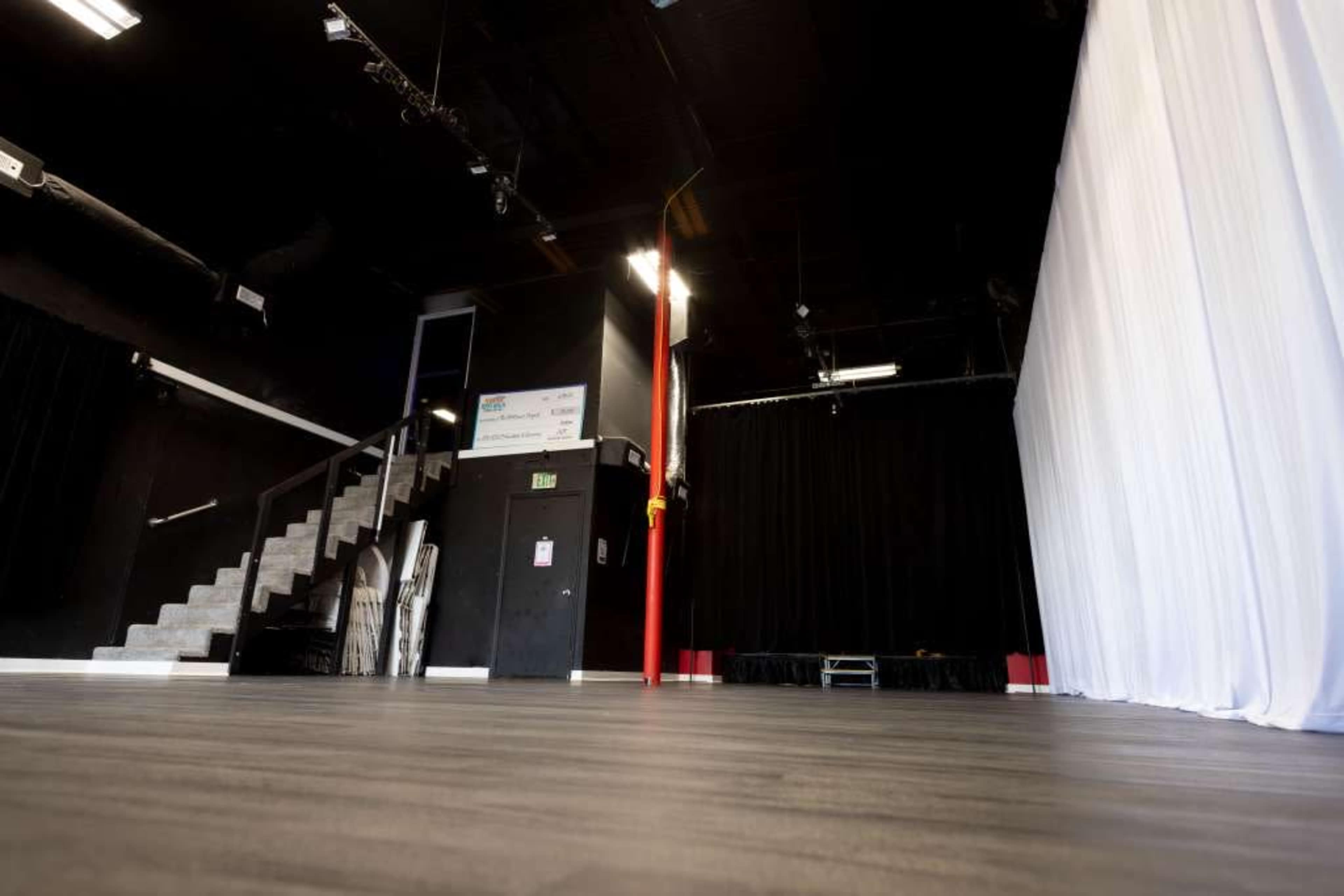 The image shows a spacious interior of a performance venue with a wooden floor, black walls, a staircase leading to the upper level, and a large white curtain on one side.