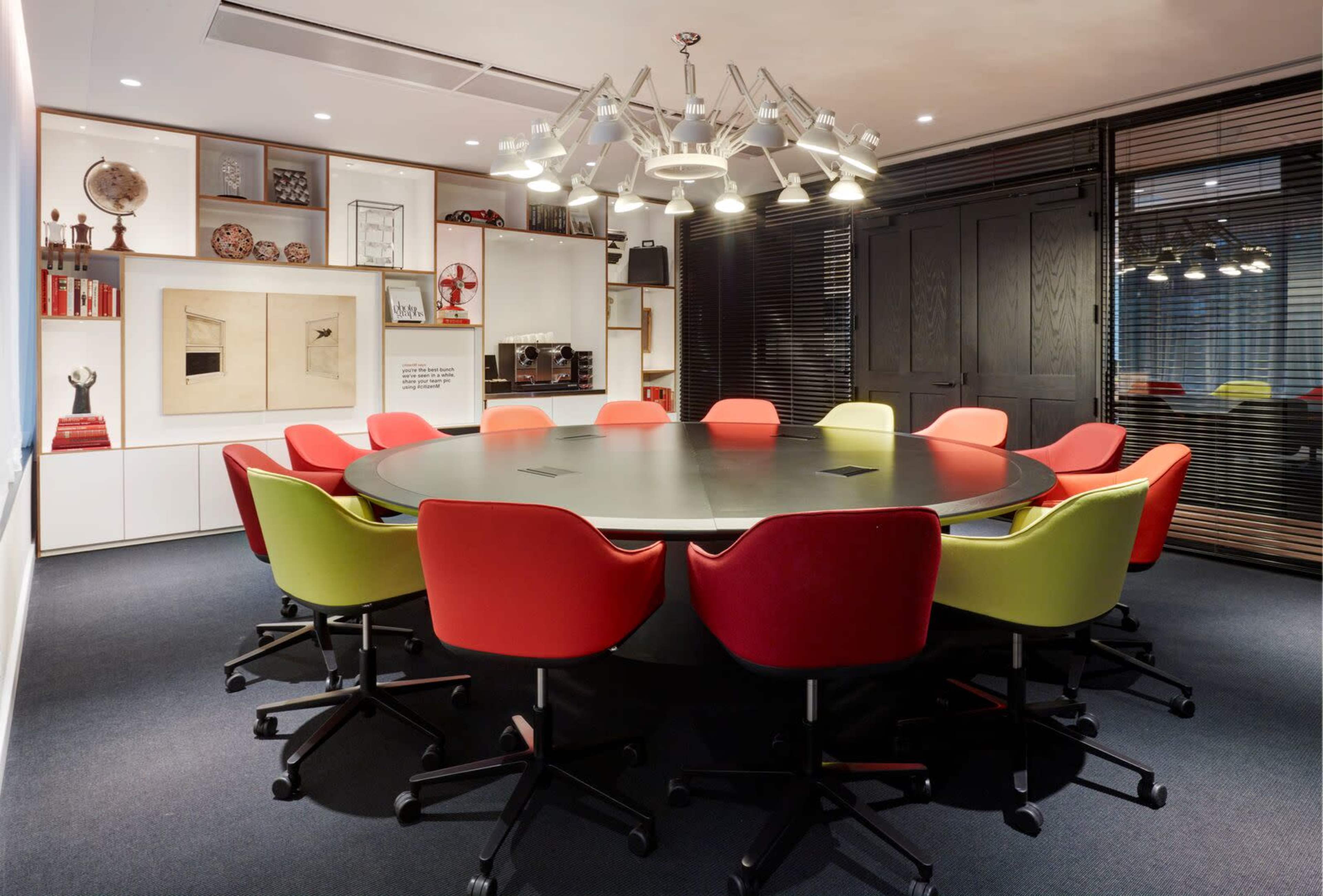 A circular conference table surrounded by colorful chairs is set in a modern meeting room with large windows and artistic decor.