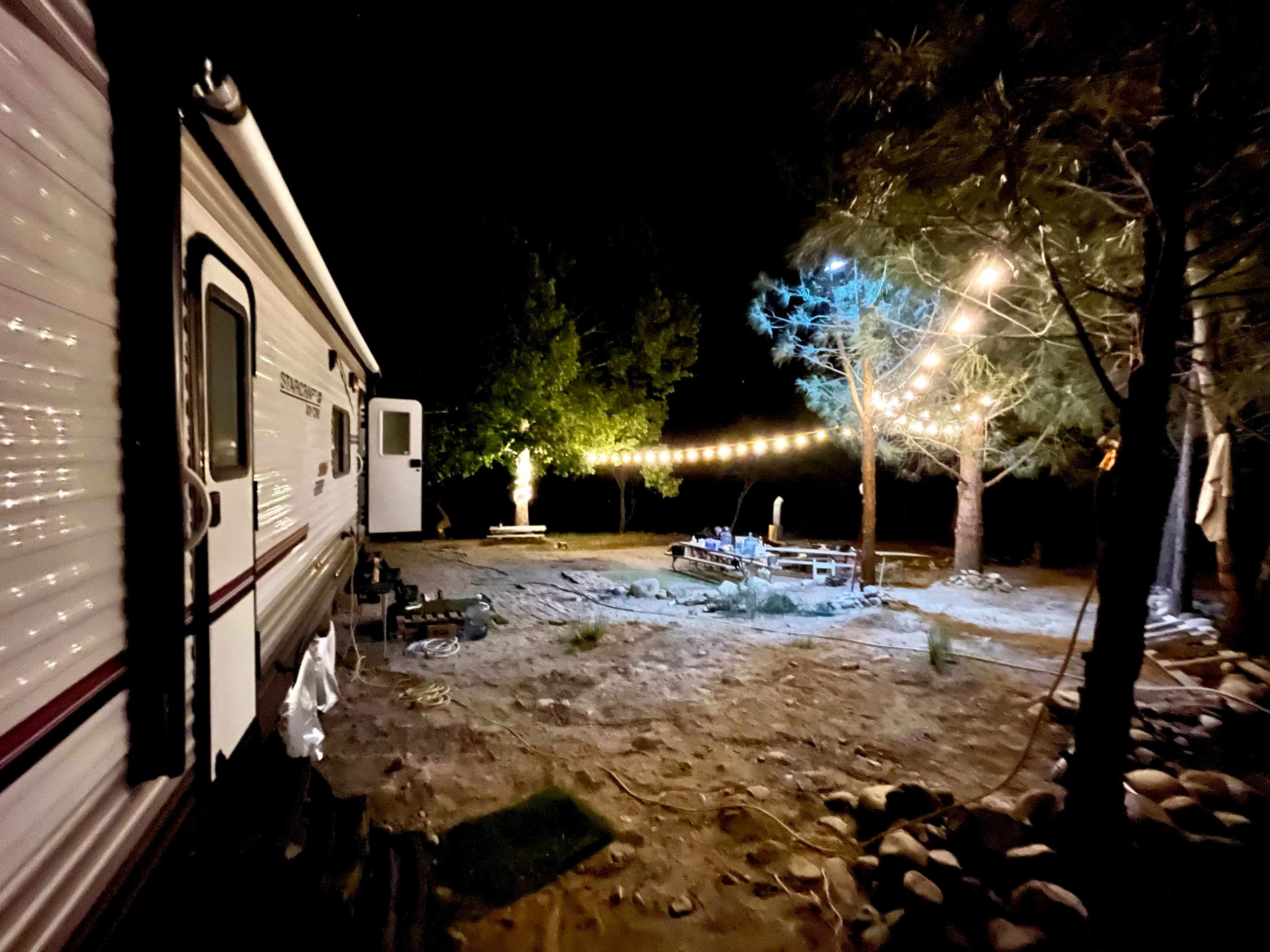 A trailer is parked next to a campsite illuminated by string lights, surrounded by trees and sandy ground.