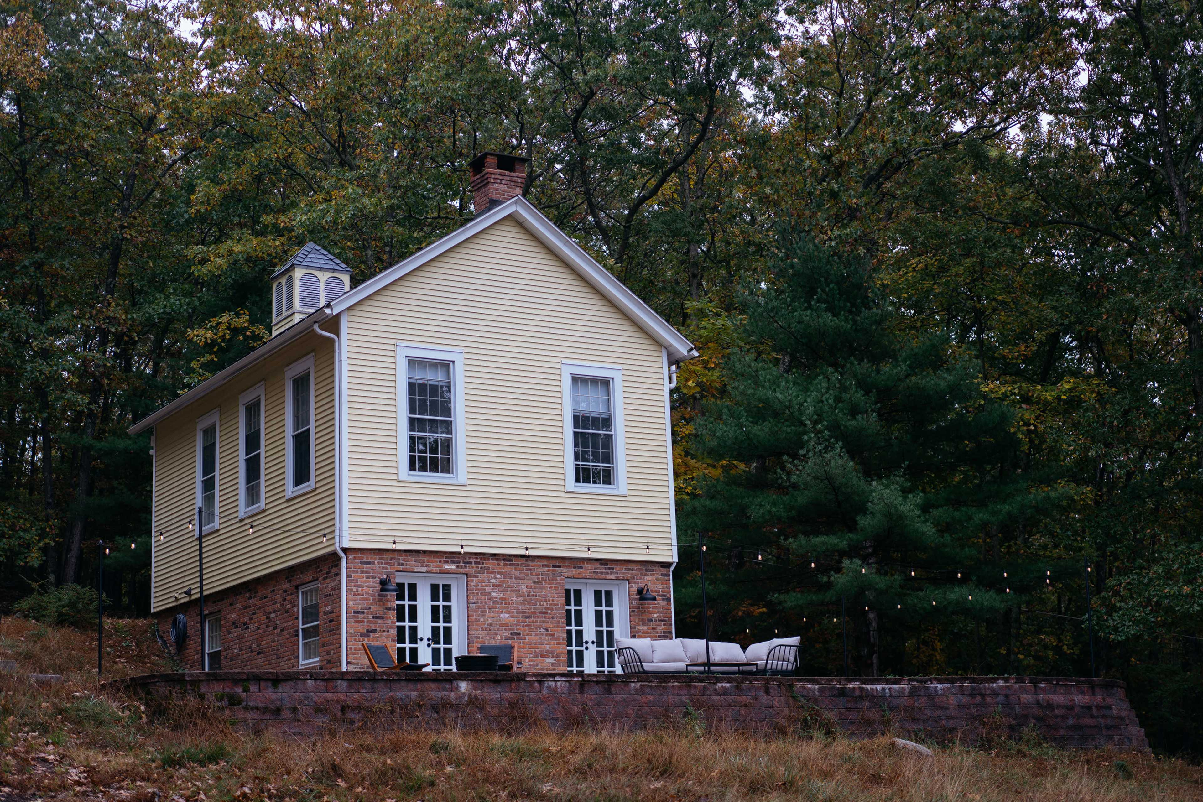 A two-story yellow house with brick accents stands surrounded by trees, featuring a patio with outdoor furniture.