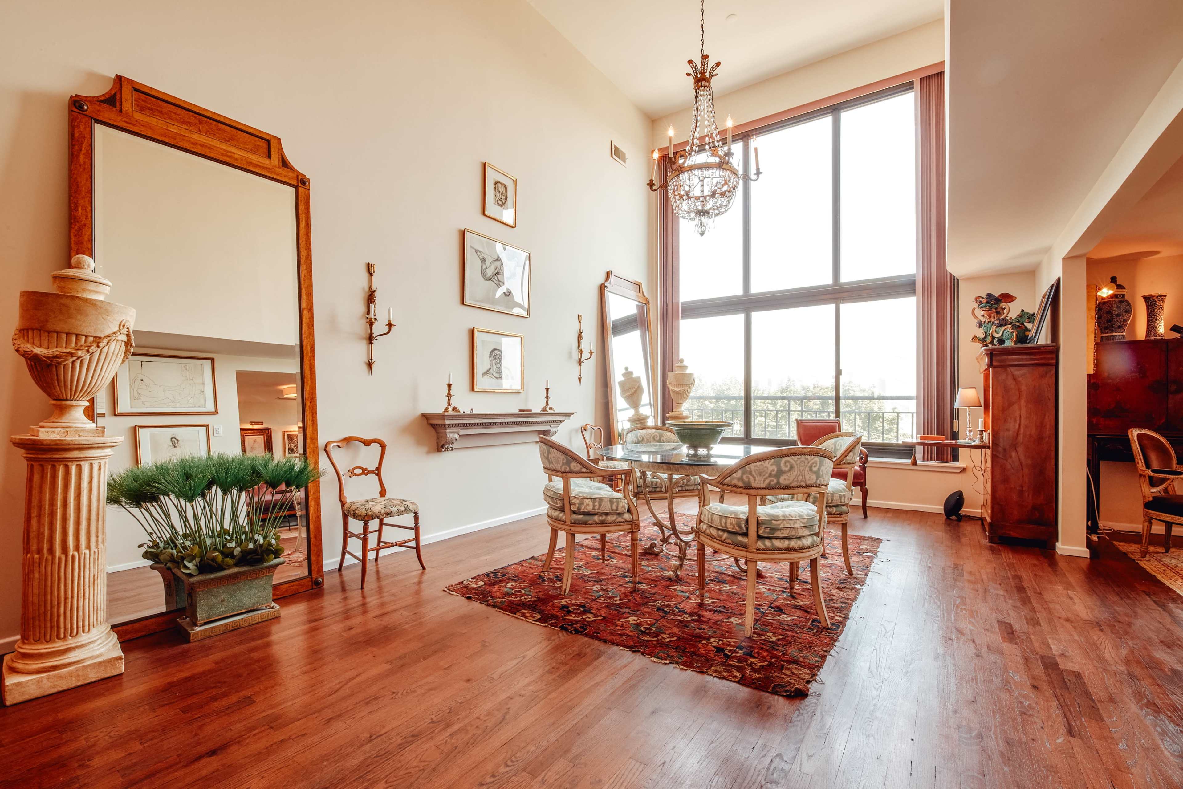 A spacious dining area with a large window, a circular glass table surrounded by upholstered chairs, and a decorative mirror along a wall.
