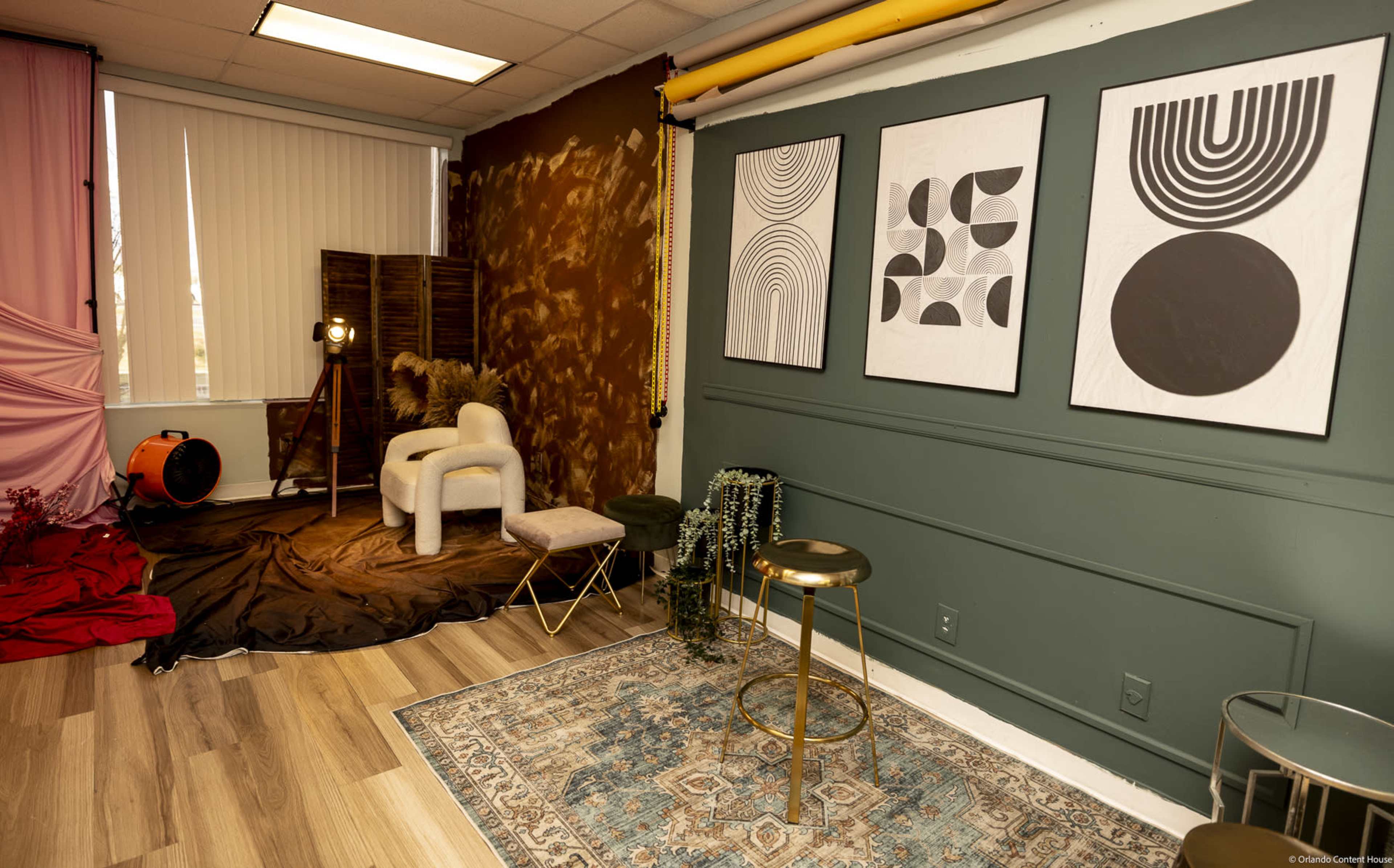 The image shows a modern interior space with abstract wall art, a small seating area, and a professional lighting setup against textured walls.