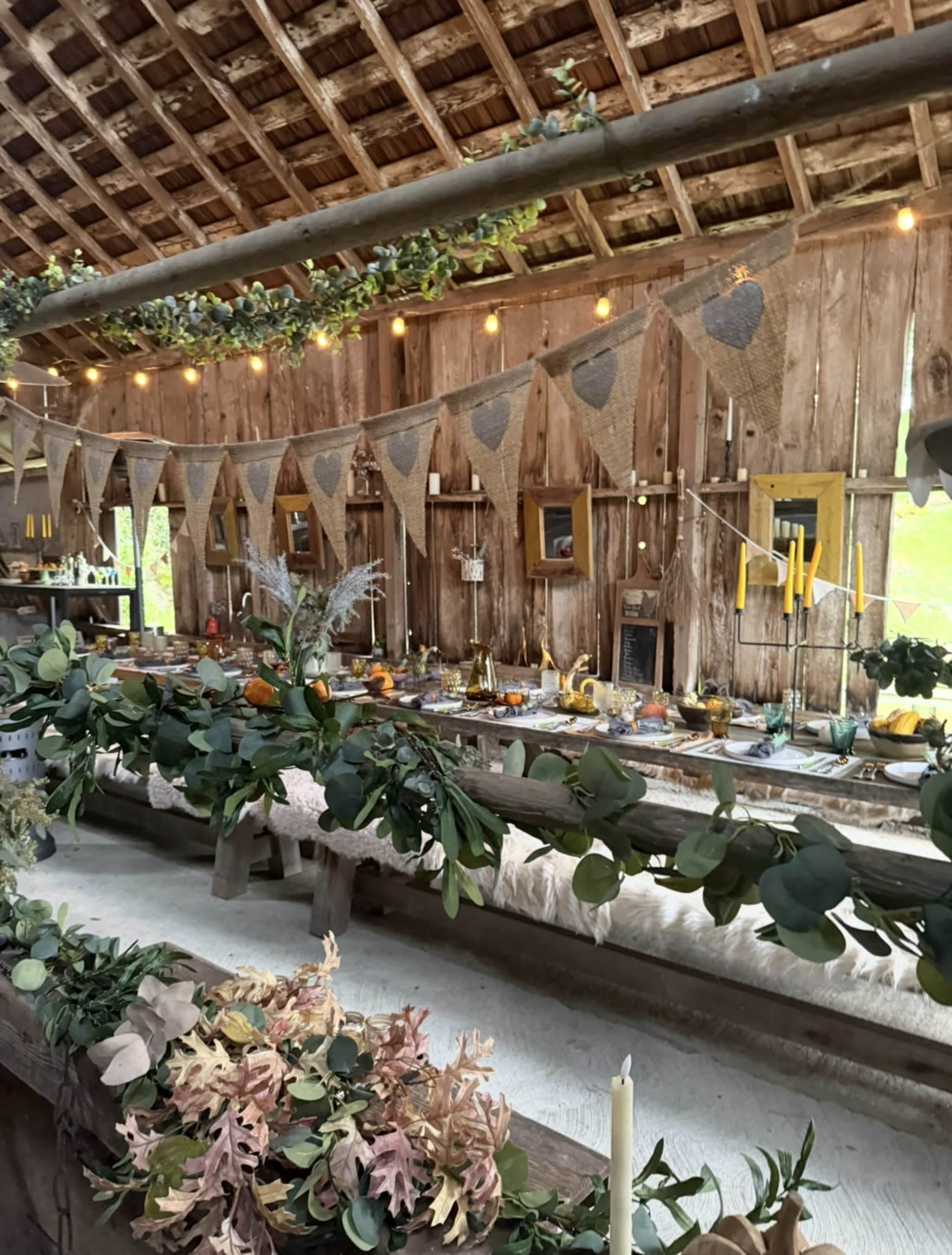 The interior of a rustic barn is decorated with garlands, bunting, candles, and a long dining table set for a gathering.