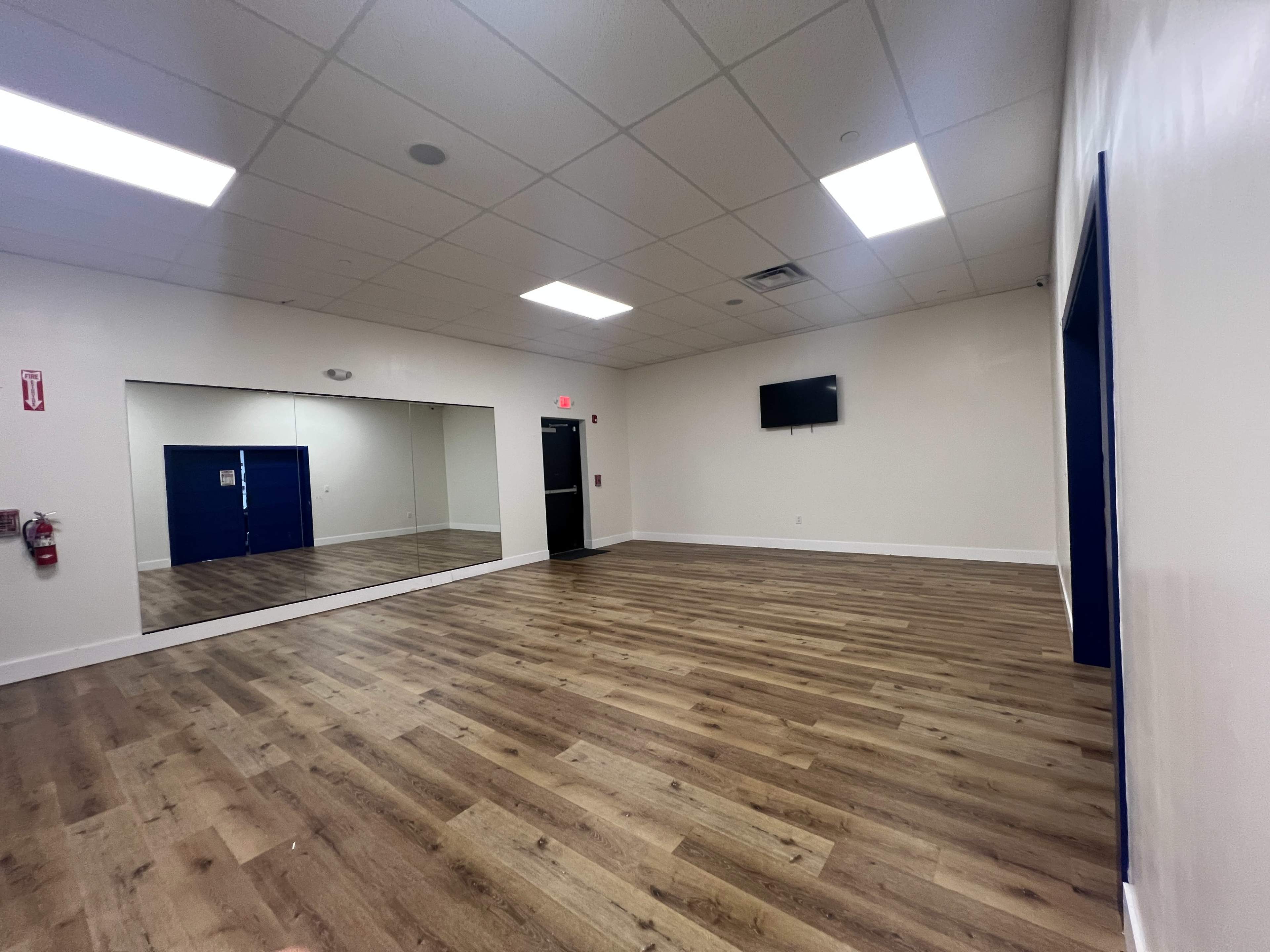 The image shows a spacious, well-lit room with wooden flooring, a mirror on one wall, and a wall-mounted television.