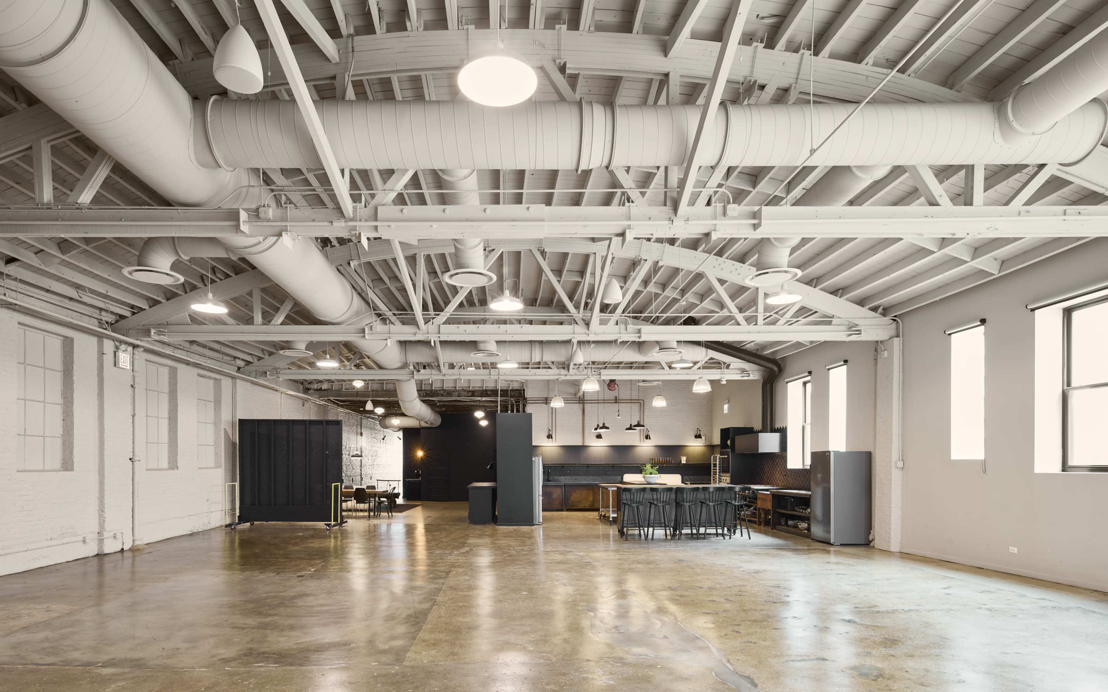 The image shows a spacious industrial-style interior with exposed wooden beams, large windows, and a kitchen area featuring a dining table.