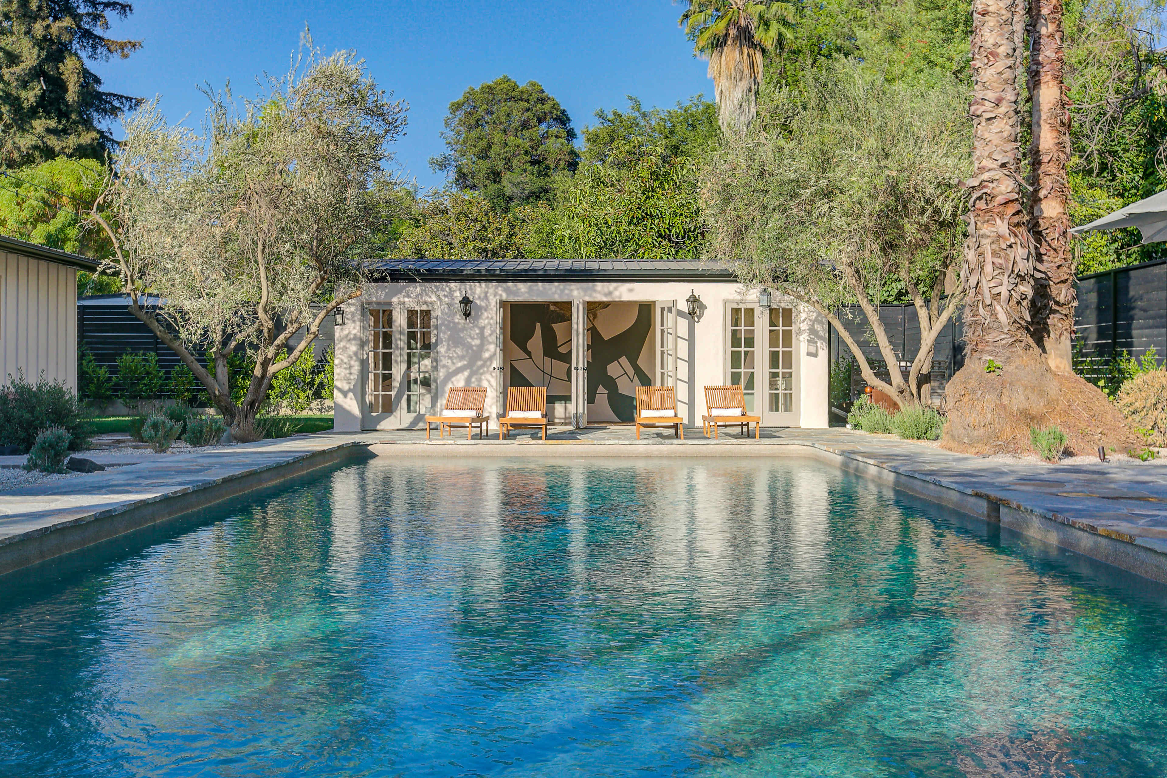 A rectangular swimming pool reflects a small building surrounded by palm and olive trees.