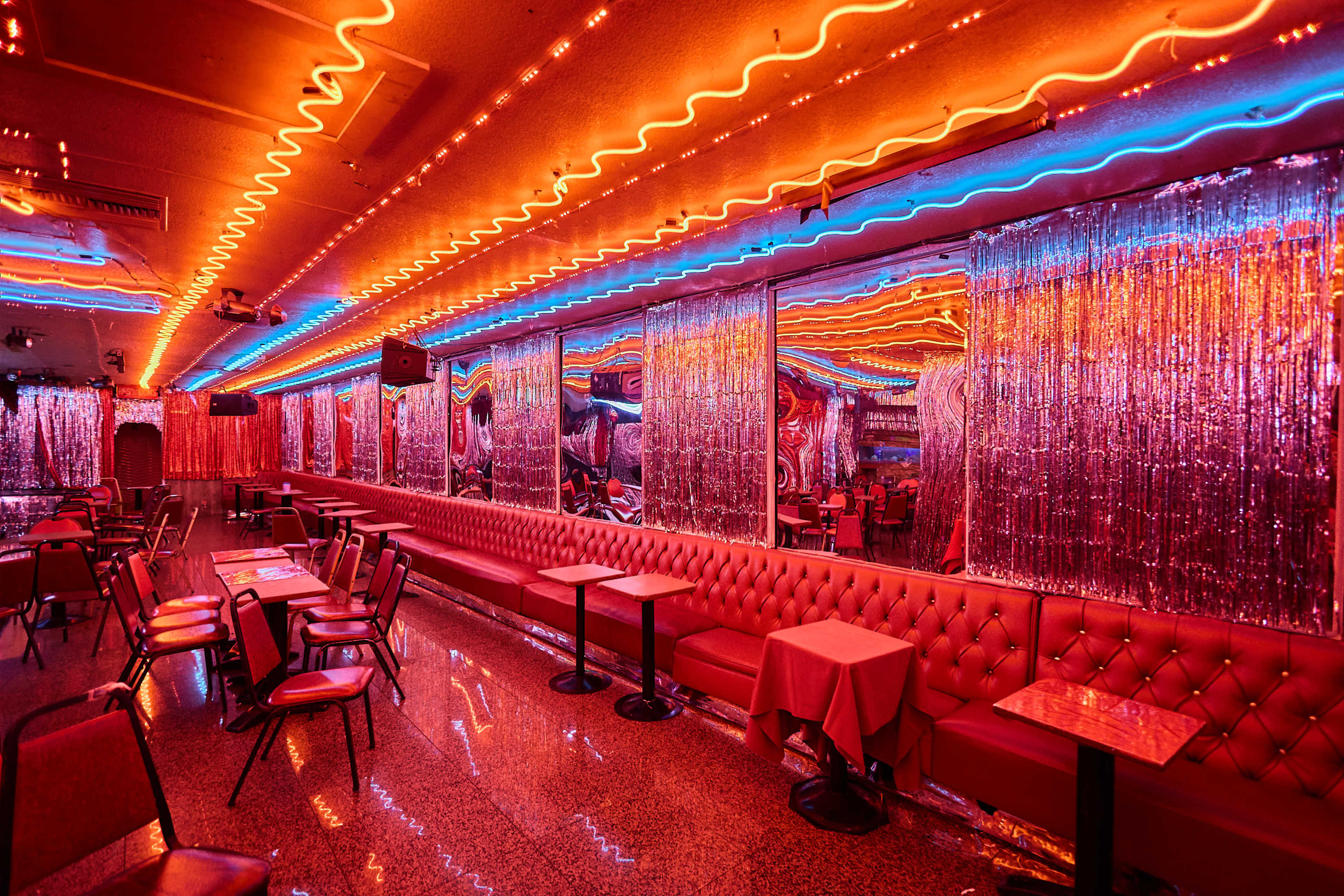 The image shows a brightly lit retro-style bar with neon lighting and mirrored walls, featuring red vinyl seating and tables arranged throughout the space.