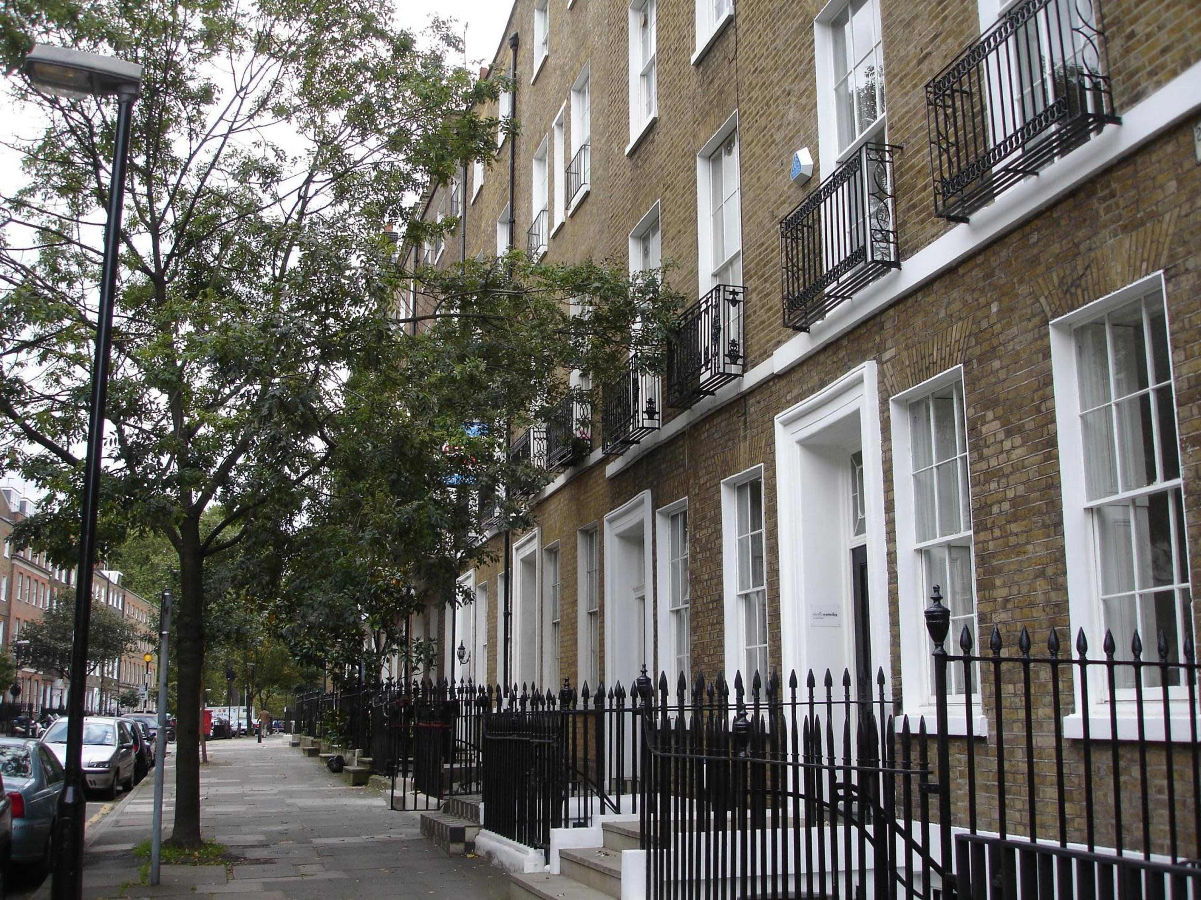 The image shows a row of brick townhouses with white trim, featuring balconies and surrounded by trees along a city street.