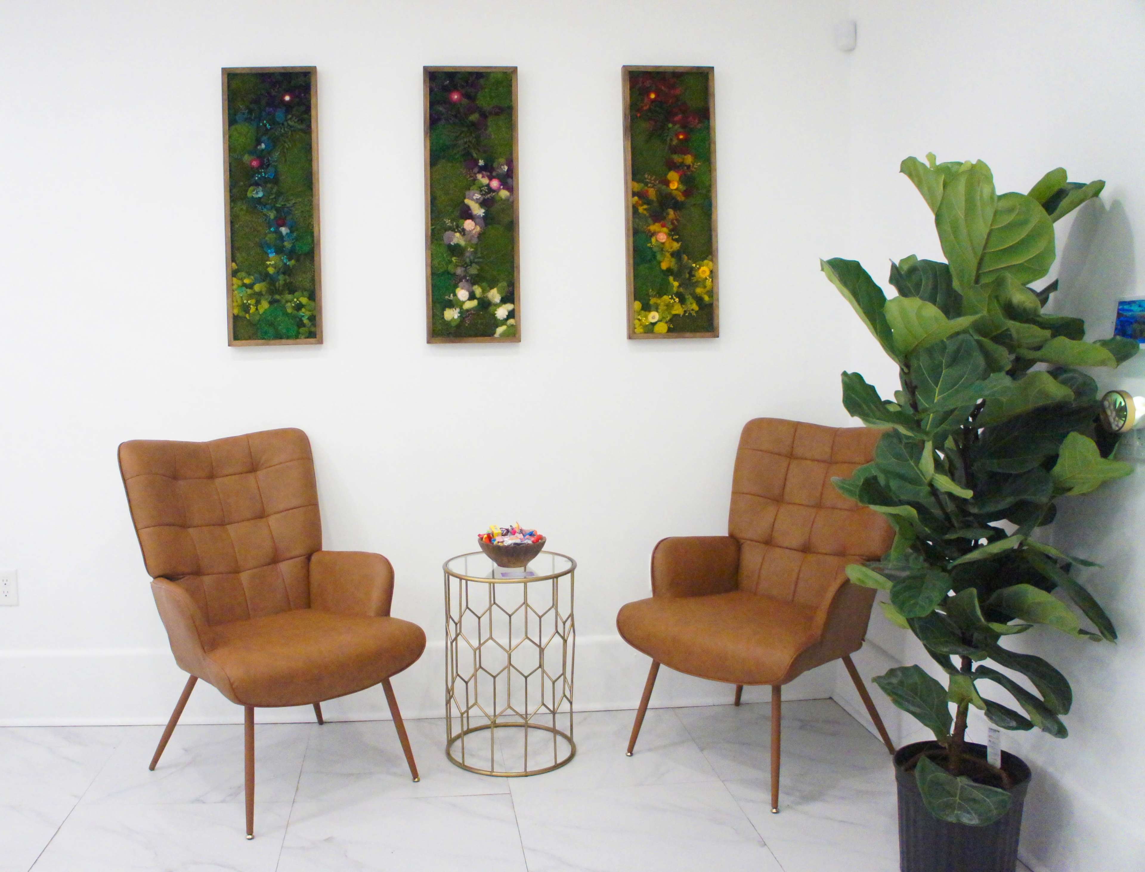 Two brown upholstered chairs sit opposite each other with a gold geometric side table between them, alongside a large potted plant and three framed pieces of floral wall art.