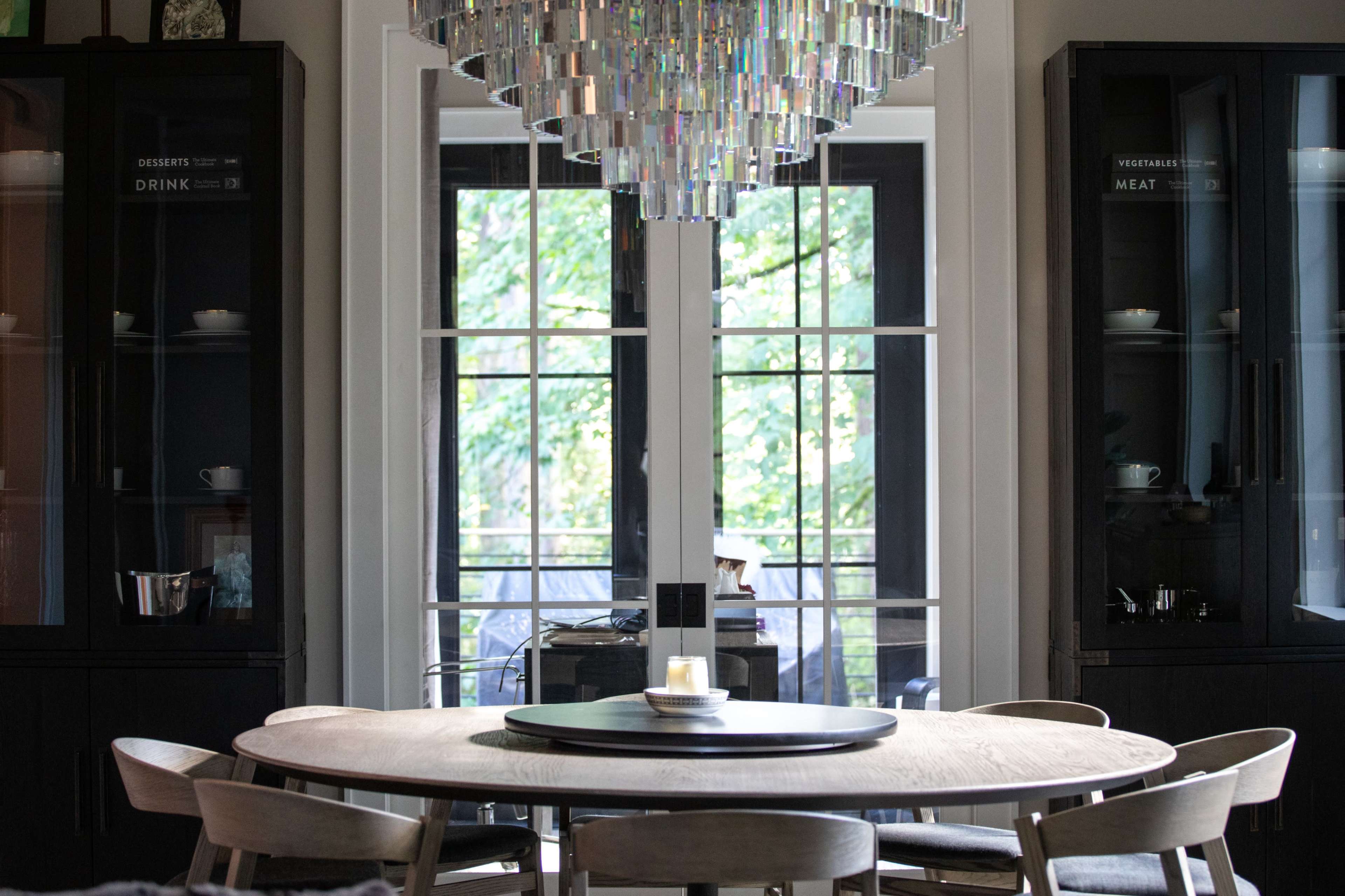 A round wooden dining table sits under a modern chandelier, surrounded by chairs, with large windows revealing greenery outside.