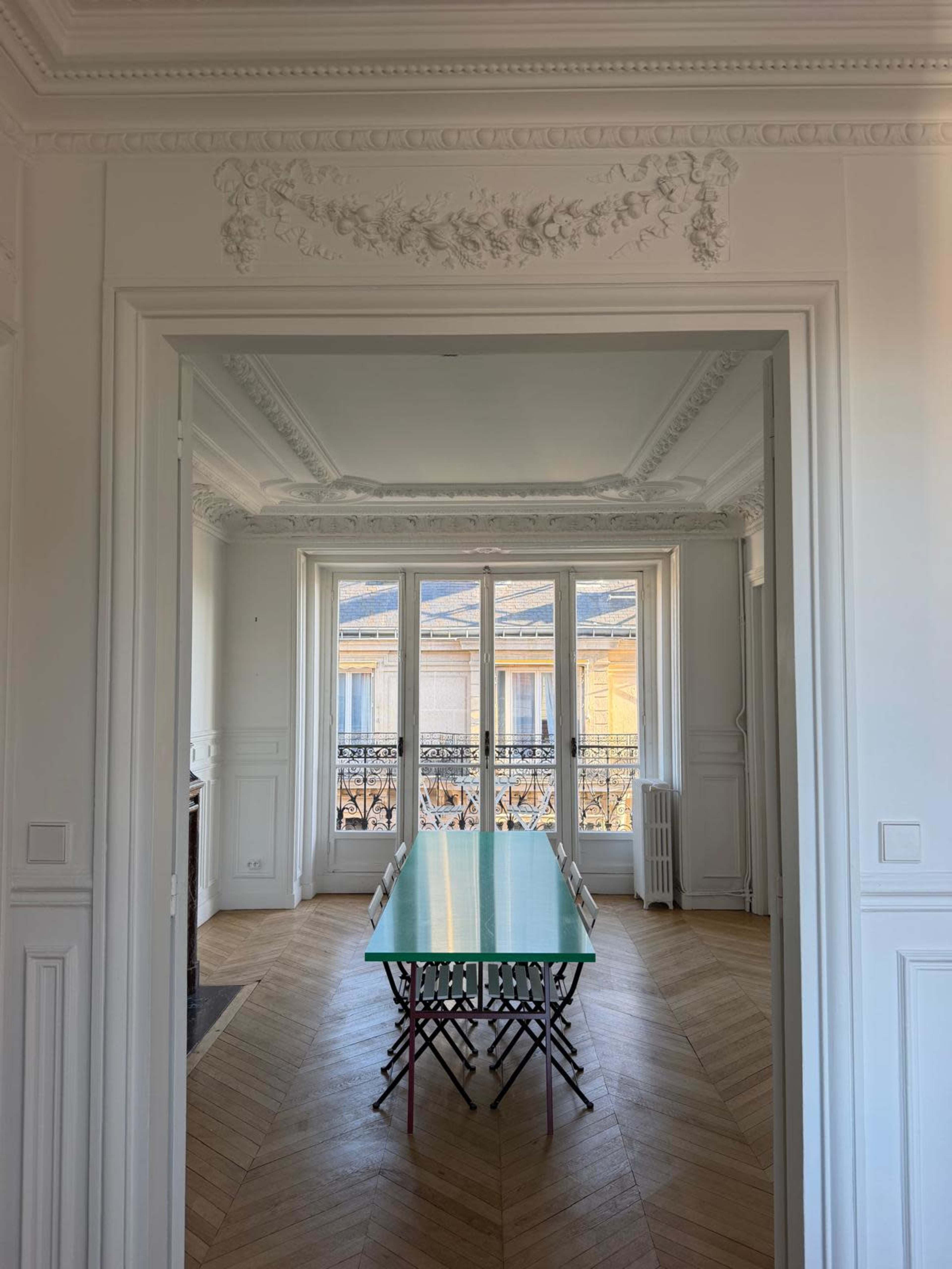 A long, green table is positioned centrally in a bright room featuring white walls and ornate moldings, with large windows overlooking a balcony.