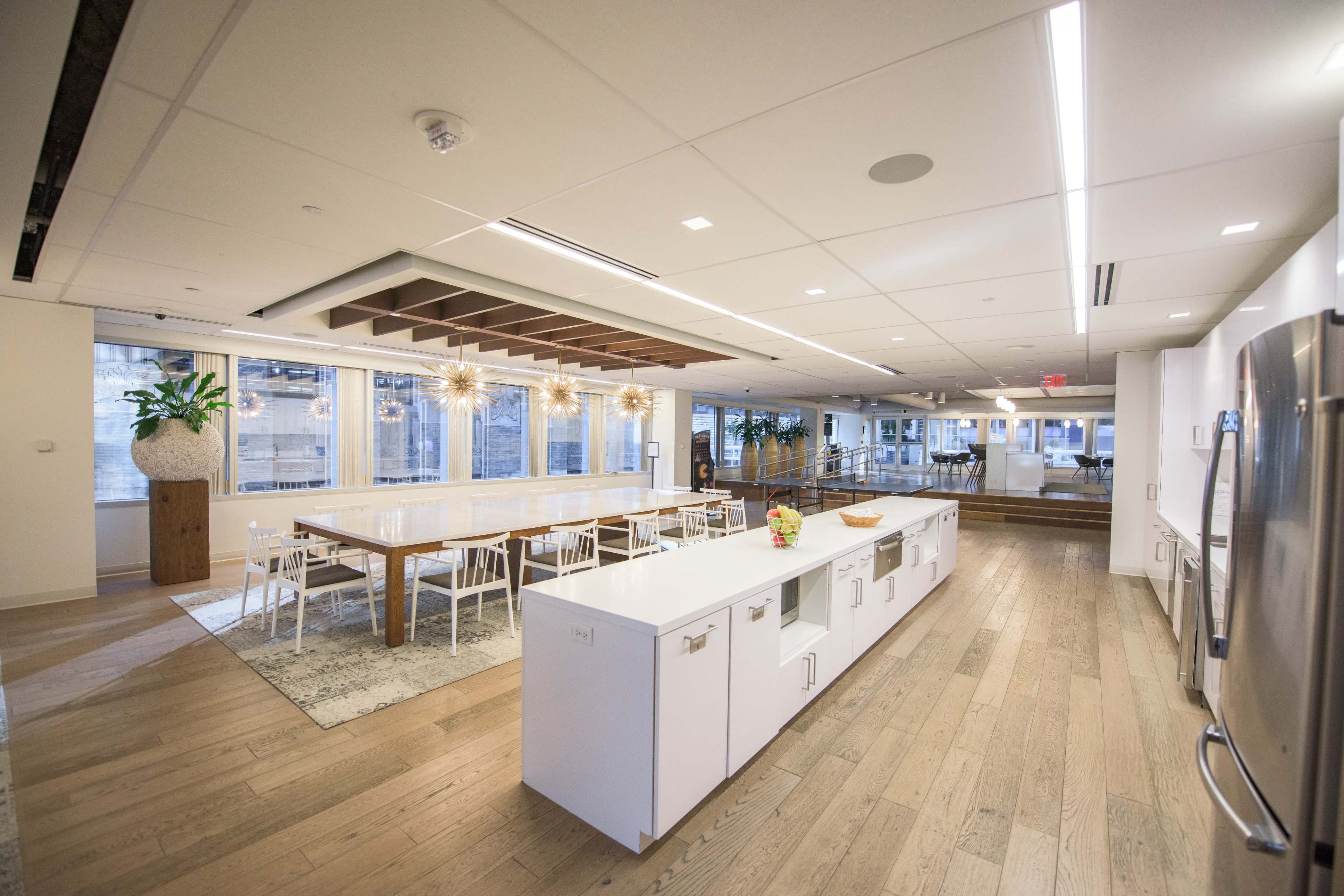 The image shows a modern office kitchen and dining area with a long white table, surrounding chairs, and large windows letting in natural light.