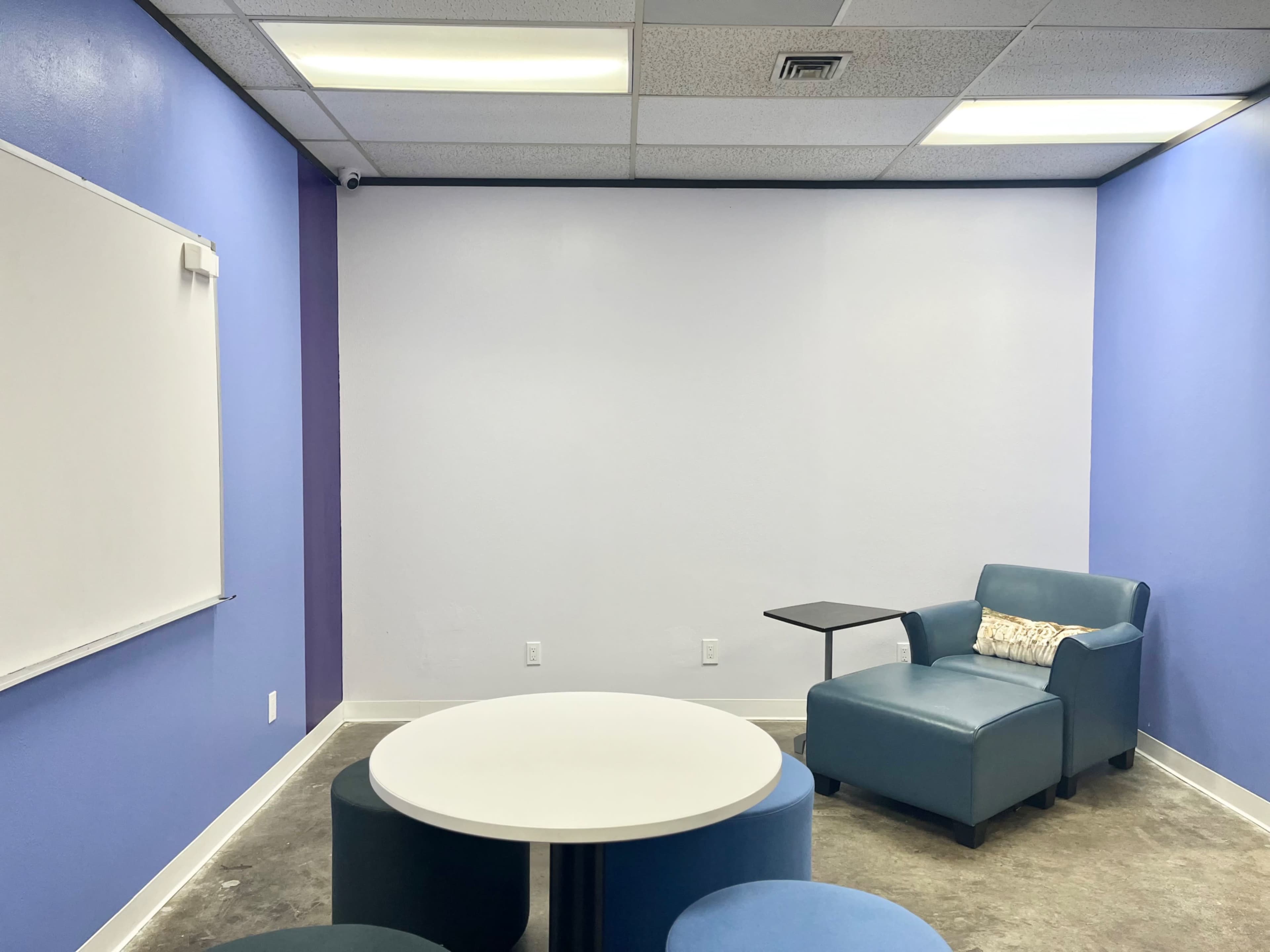The image shows a small room with light blue walls, a whiteboard, a round white table, and a blue armchair with a side table.